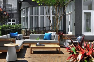 Un área de estar al aire libre con sofá, mesa de centro y sillas, rodeada de plantas y un edificio con grandes ventanales.
