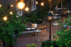 An outdoor seating area with tables and benches, surrounded by greenery and illuminated by string lights. - Kimpton Brice Hotel by IHG (Savannah)