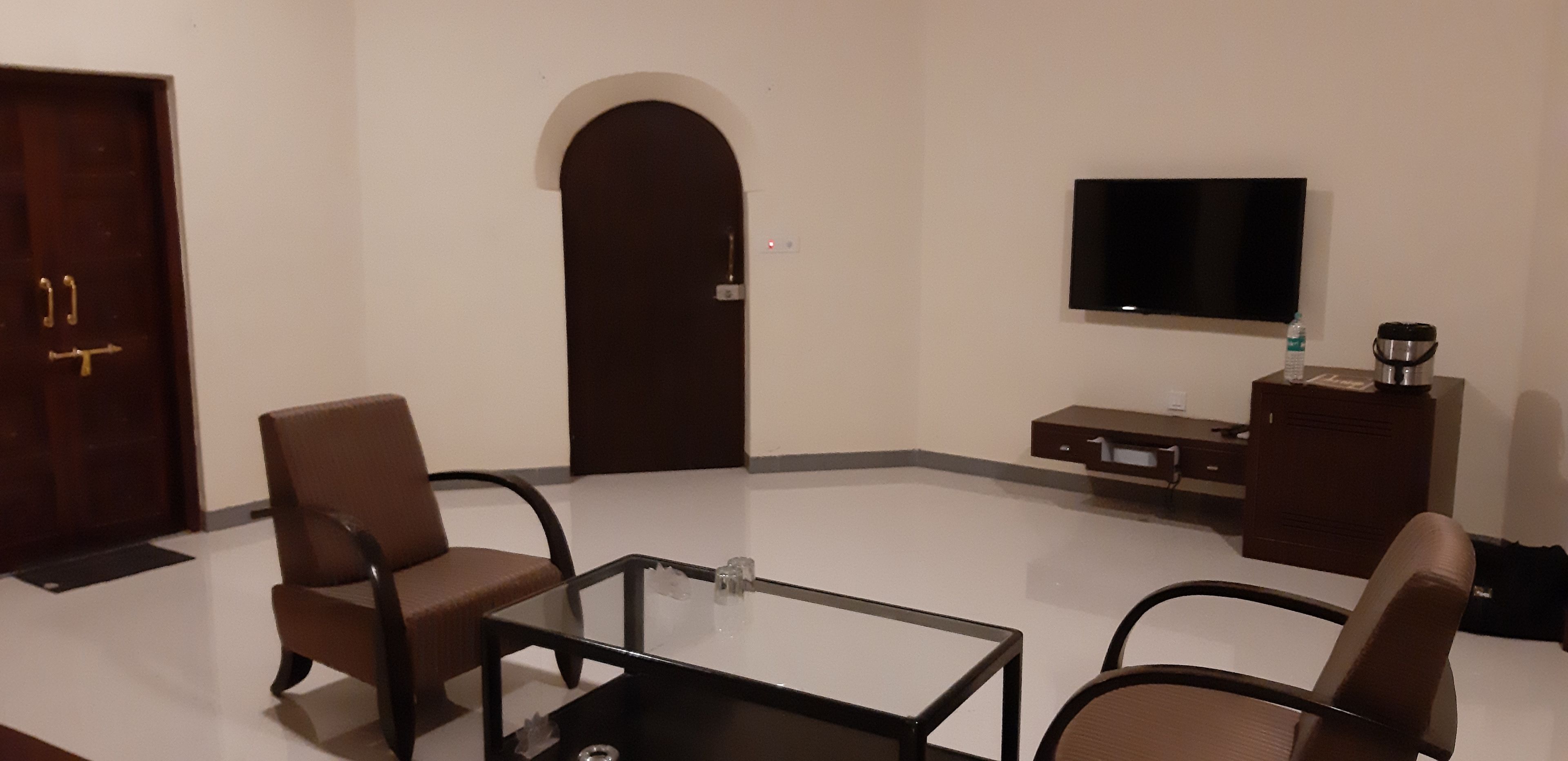 Habitación de hotel con una mesita de vidrio, dos sillas color café, un televisor de pantalla plana, un minibar y una puerta con cerradura electrónica.