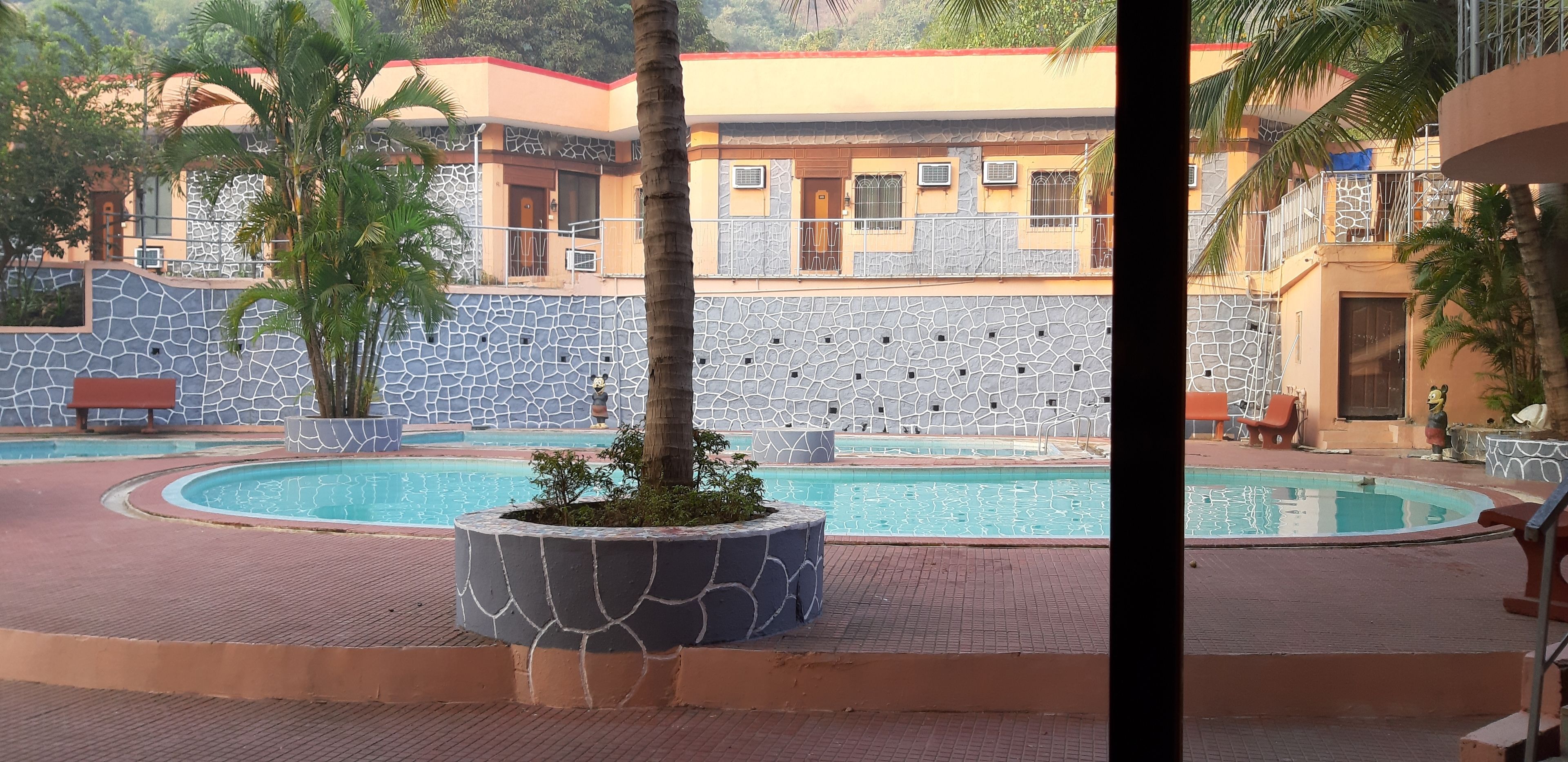 A swimming pool with a stone wall and palm trees, surrounded by a building with a red roof and a stone facade.