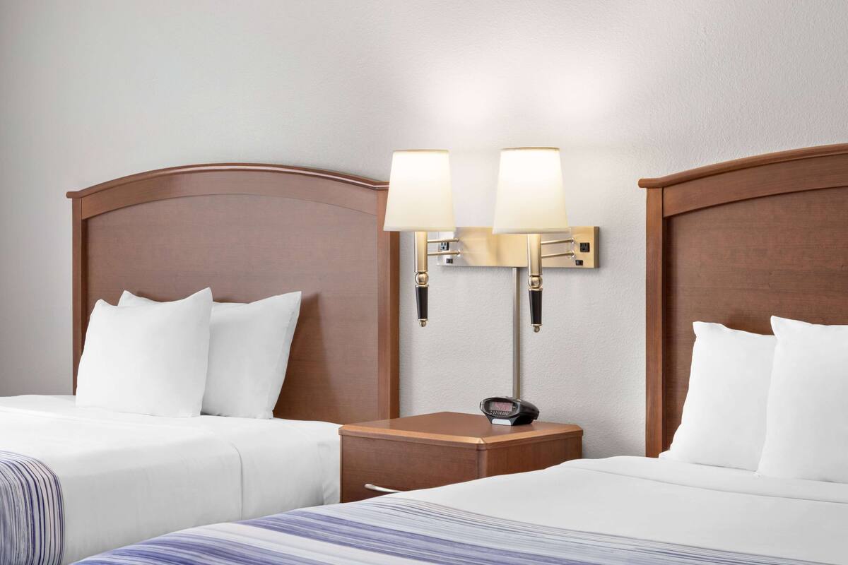 Ein Hotelzimmer mit zwei Betten, jeweils mit weißer Bettwäsche und einem hölzernen Kopfteil. Zwischen den Betten steht ein Nachttisch mit einer Lampe und einer Uhr.