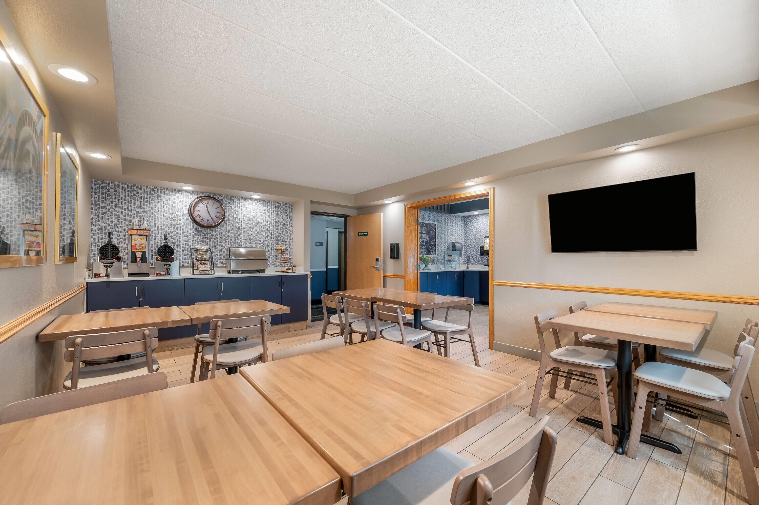 A dining area with wooden tables and chairs, a kitchen in the background, and a flat-screen TV mounted on the wall.