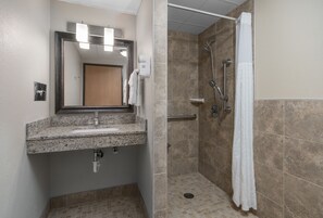 A bathroom with a granite countertop sink, a mirror, a towel rack, a shower with a glass door, and beige tiled walls. - AmericInn by Wyndham Bemidji (Bemidji)