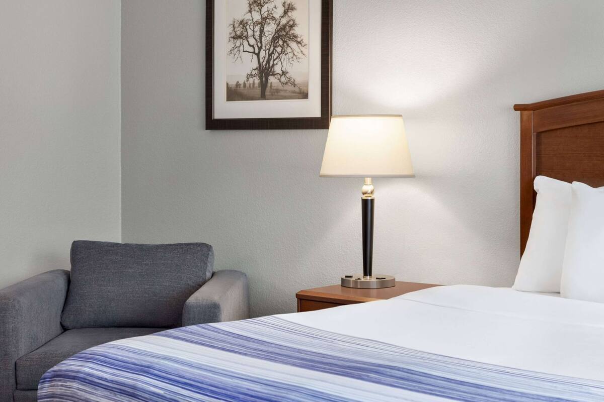 Ein Hotelzimmer mit einem Bett, einem Sessel, einem Nachttisch mit Lampe sowie einem gerahmten Bild an der Wand.