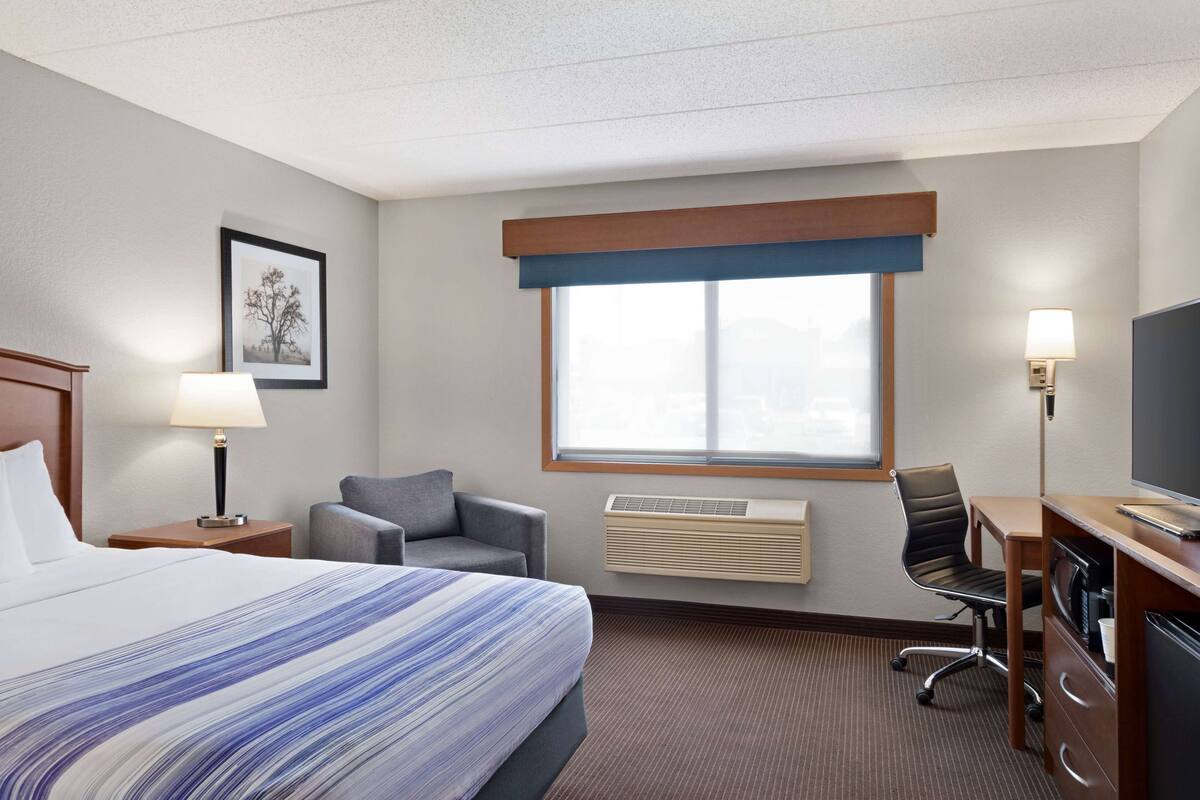 Ein Hotelzimmer mit Bett, Schreibtisch, Stuhl, Fernseher, einem Bild an der Wand und einer Klimaanlage.