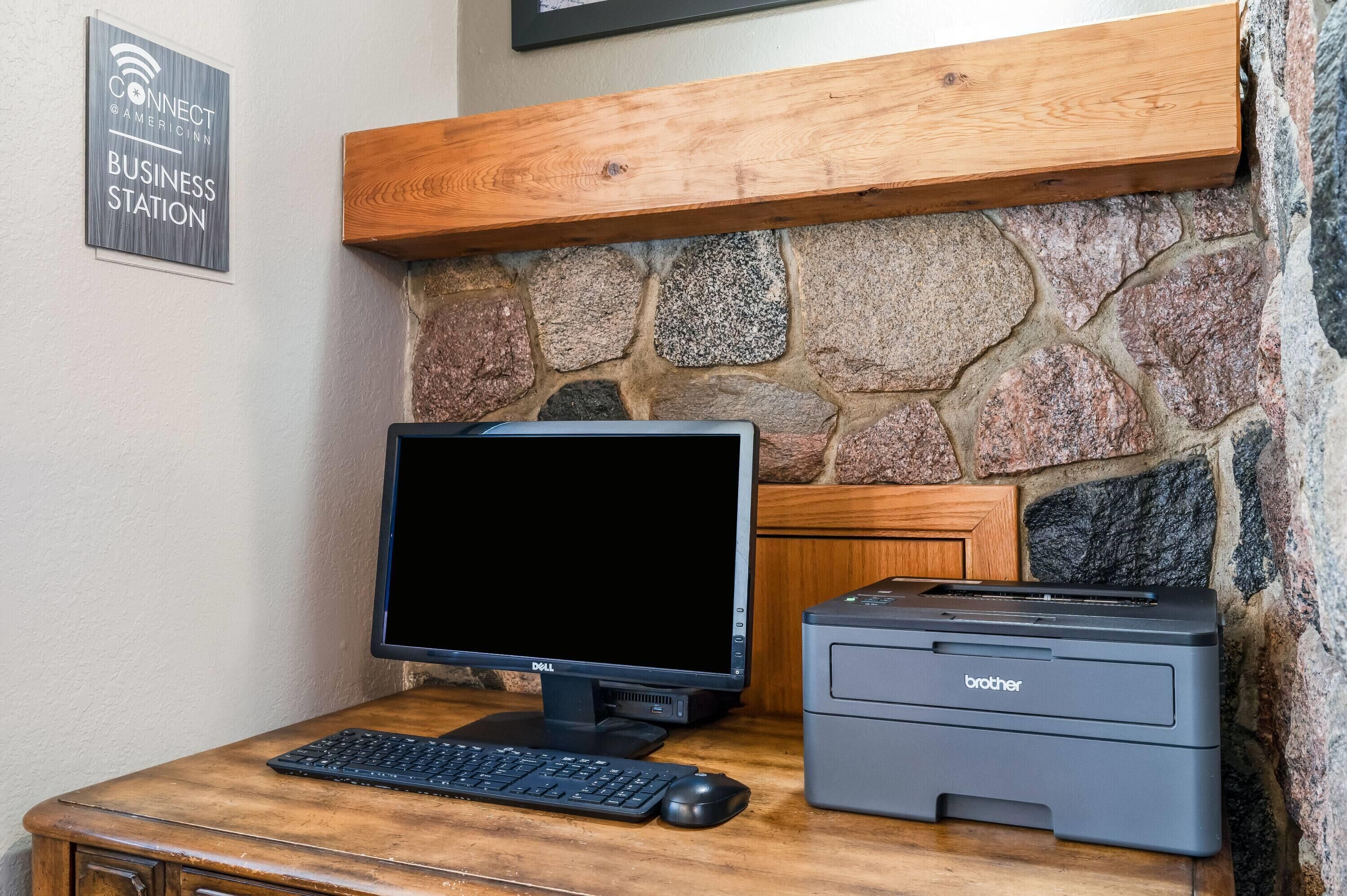 Een houten bureau met een Dell monitor en een Brother printer, een stenen muur, en een bordje met de tekst 'CONNECT @AMERICINN BUSINESS STATION'.