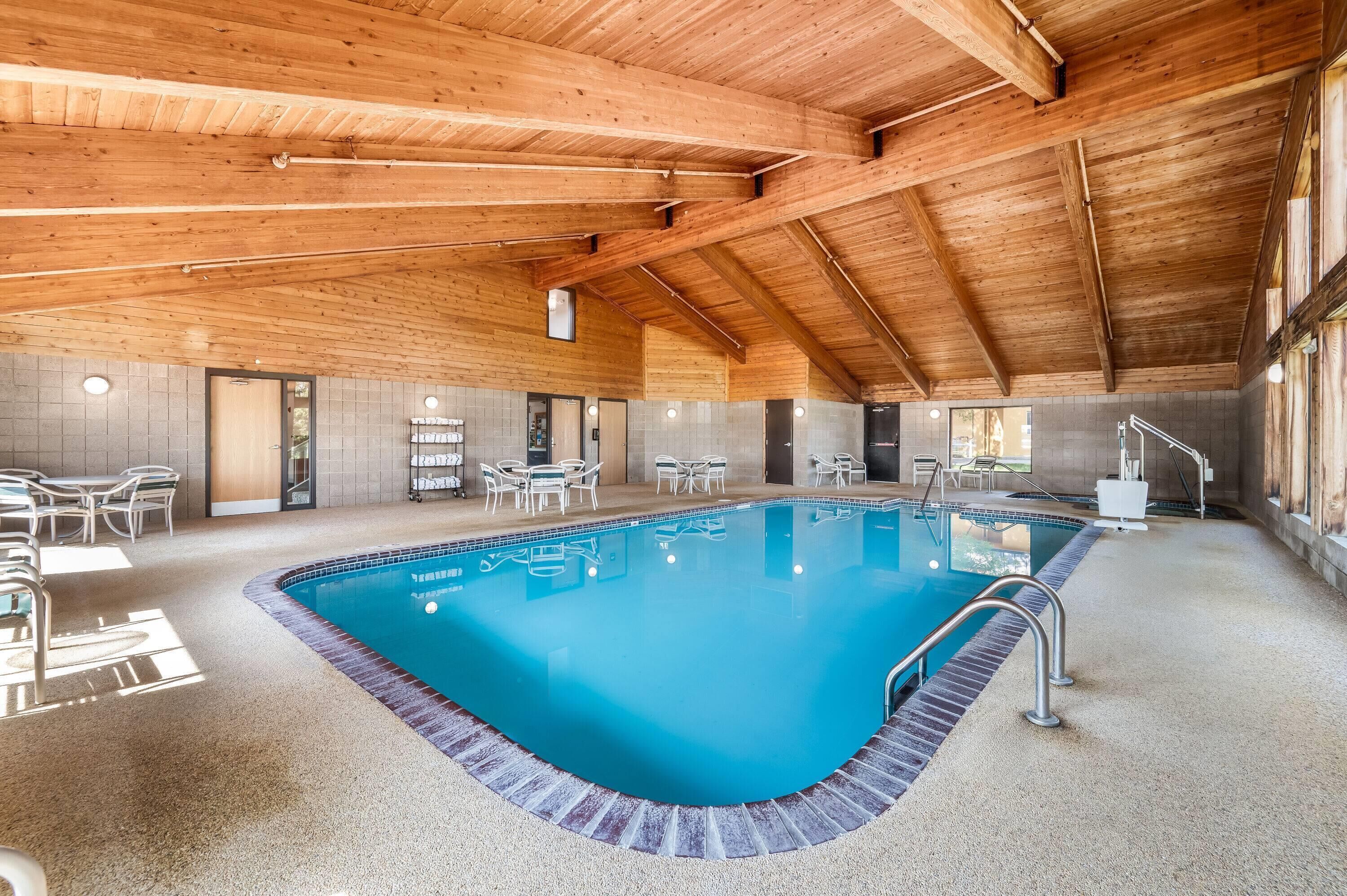 Una piscina interior con piso de baldosas, rodeada de sillas y mesas blancas, bajo un techo de madera.