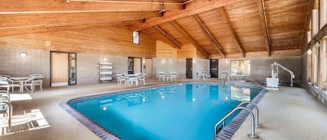 An indoor swimming pool with a tiled floor, surrounded by white chairs and tables, under a wooden ceiling.