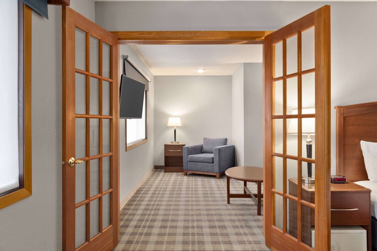 Ein Hotelzimmer mit Schachbrettmuster-Teppich, einem kleinen runden Tisch, einem Sessel, einem Nachttisch mit Lampe und einem an der Wand befestigten Fernseher.