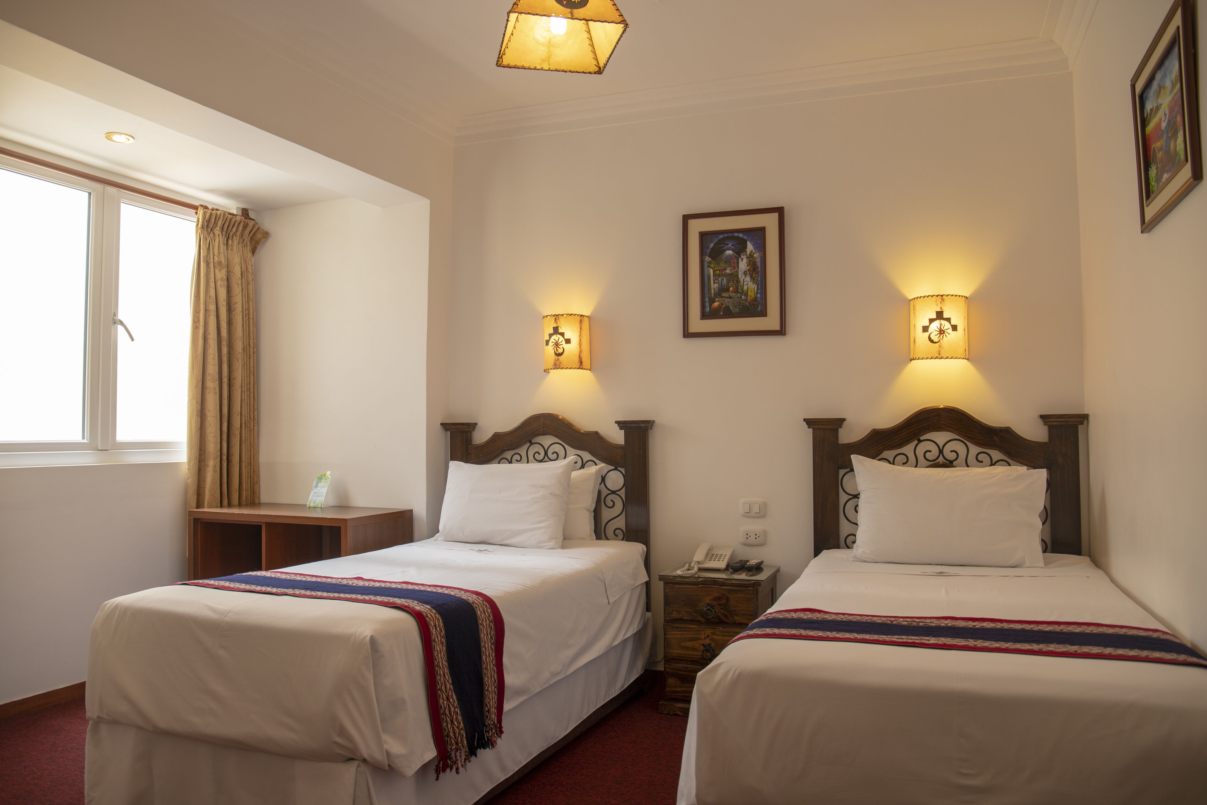A hotel room with two beds, a nightstand, a lamp, a painting, and a window with curtains.