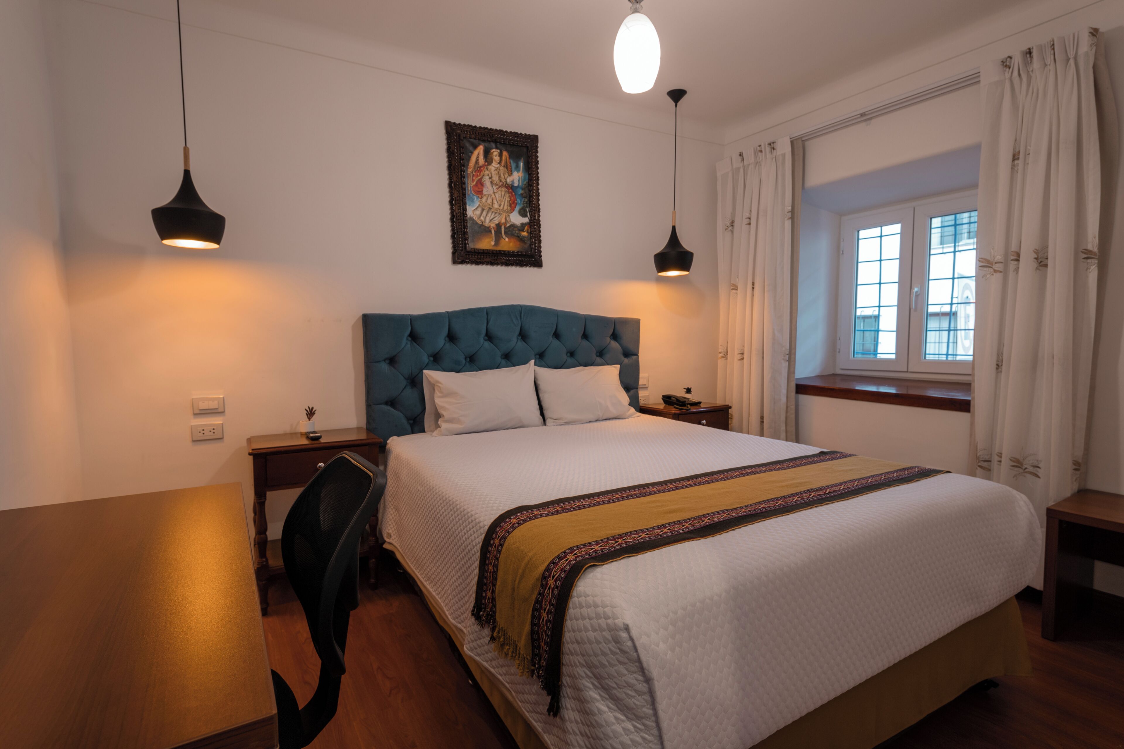 A bedroom with a large bed, a desk, two pendant lights, a nightstand, and a window with curtains.