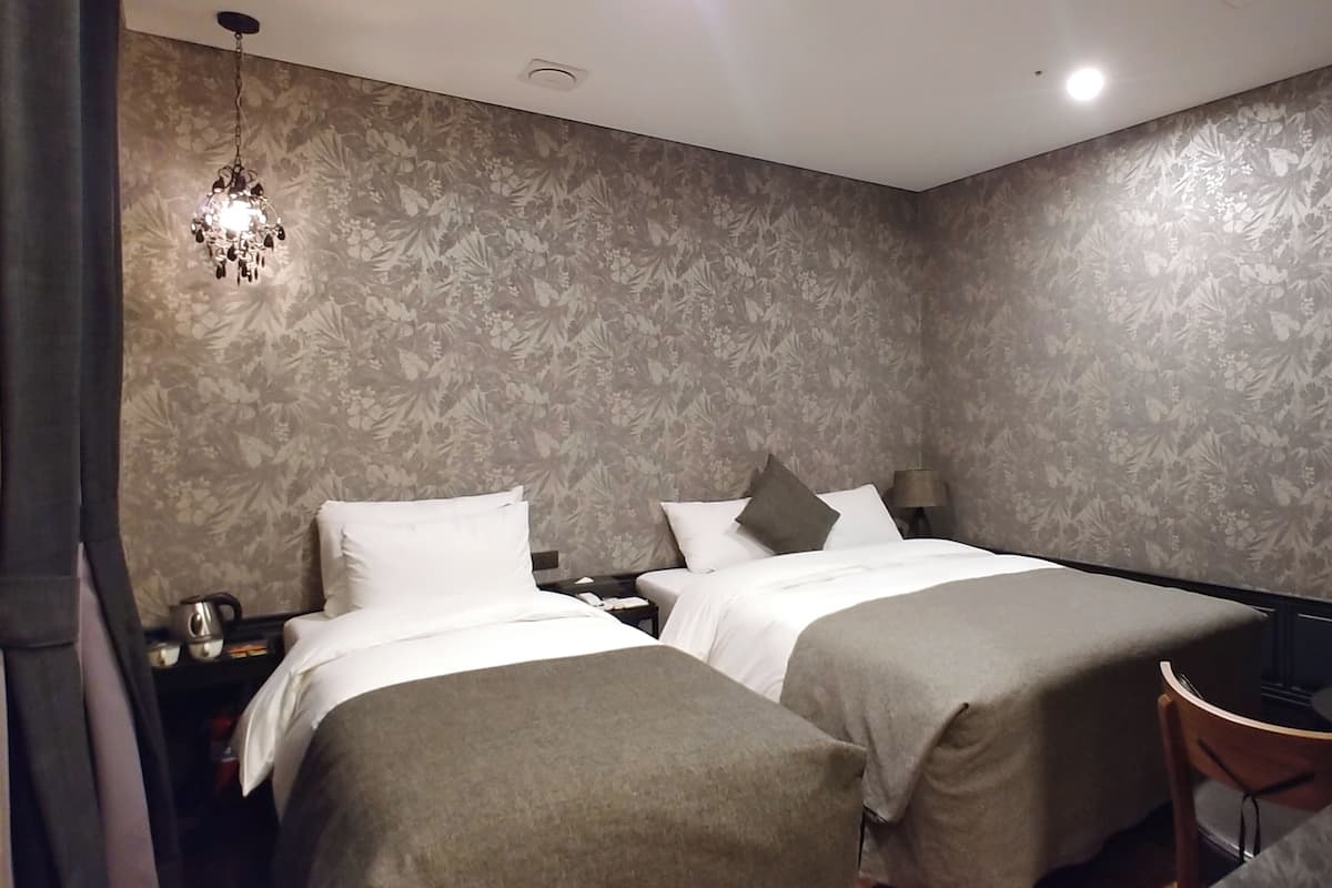 A hotel room with two beds, a nightstand, a lamp, and a chair.