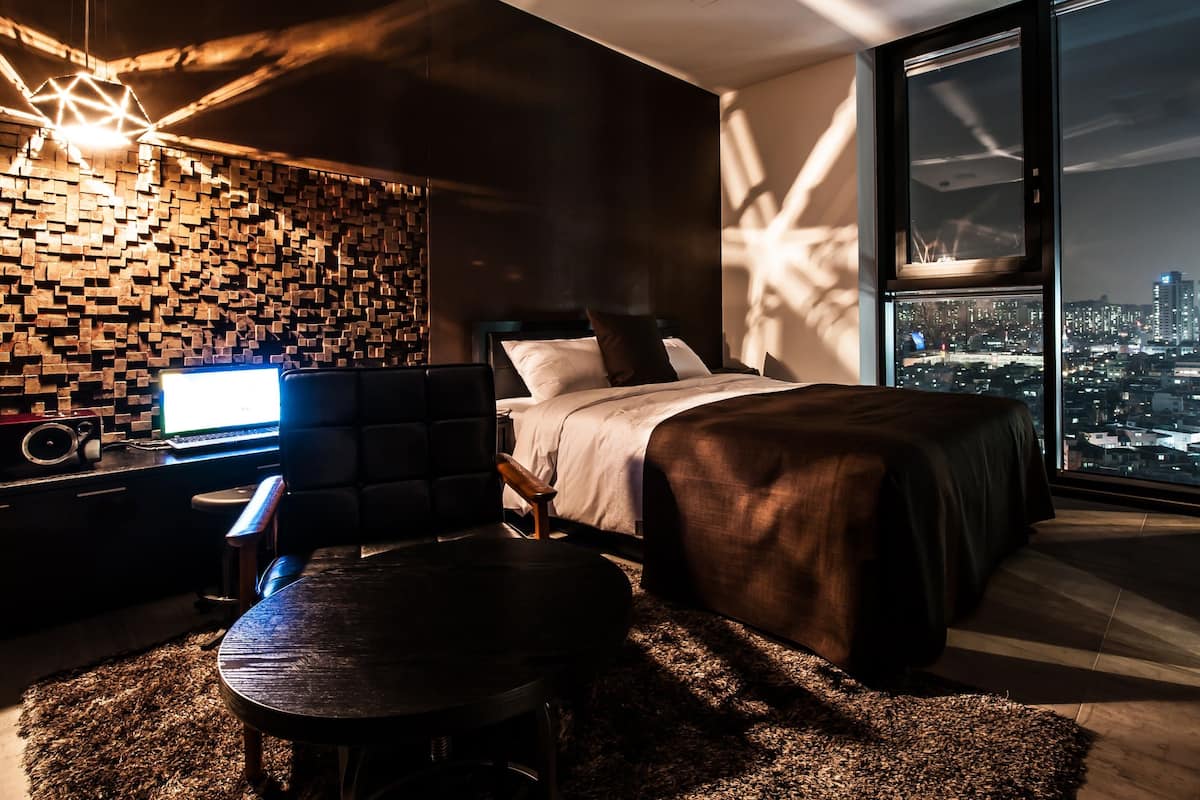 A modern bedroom with a city view, a wooden wall, a desk with a laptop, and a cozy seating area.