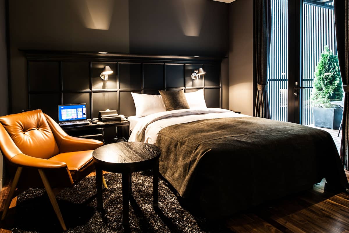 A modern bedroom with a bed, a leather chair, a small table, and a laptop on a nightstand.