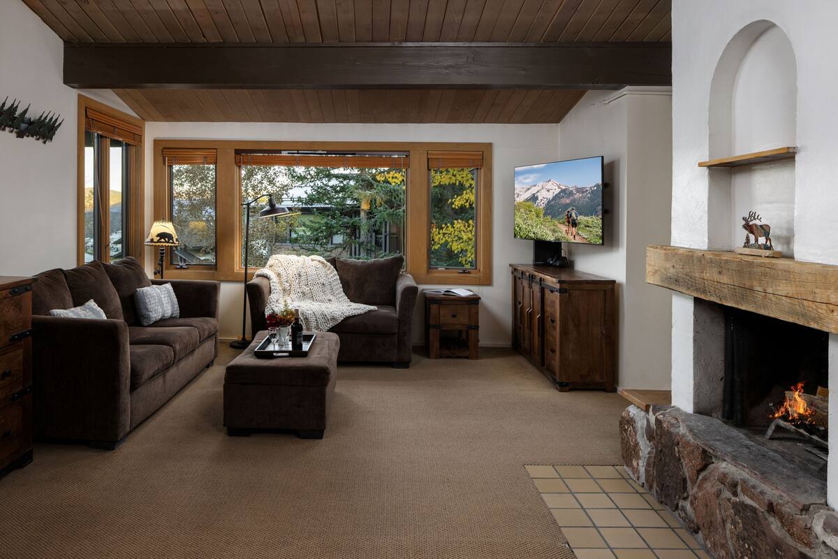 A cozy living room with a fireplace, wooden furniture, and large windows offering a view of the outdoors.