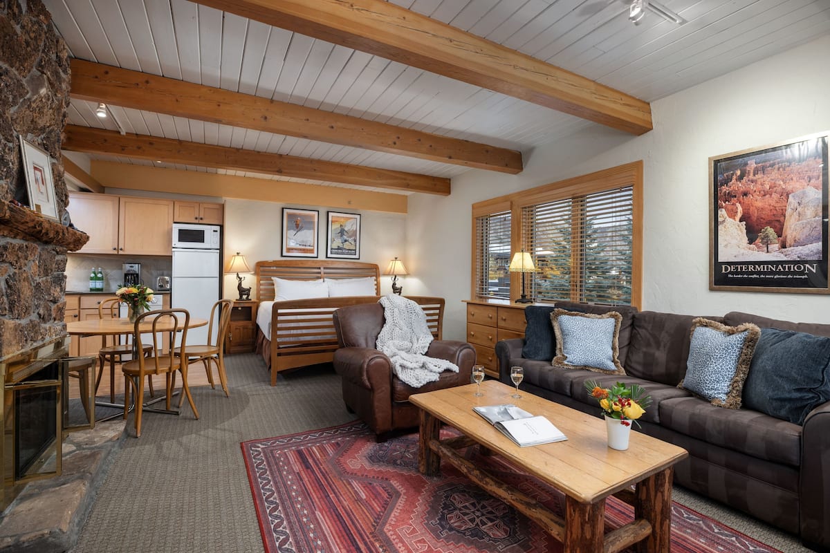 A cozy living room with a fireplace, wooden ceiling beams, a dining area with a table and chairs, and a sofa with decorative pillows.
