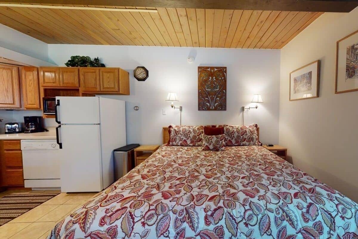 A bedroom with a patterned bedspread, a wooden ceiling, a kitchen with wooden cabinets, and a wall clock.