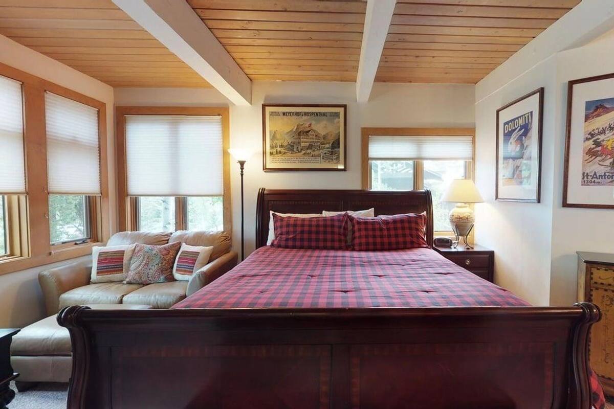 A bedroom with a wooden bed, a sofa, and a nightstand. There are framed pictures on the walls and a window with blinds.