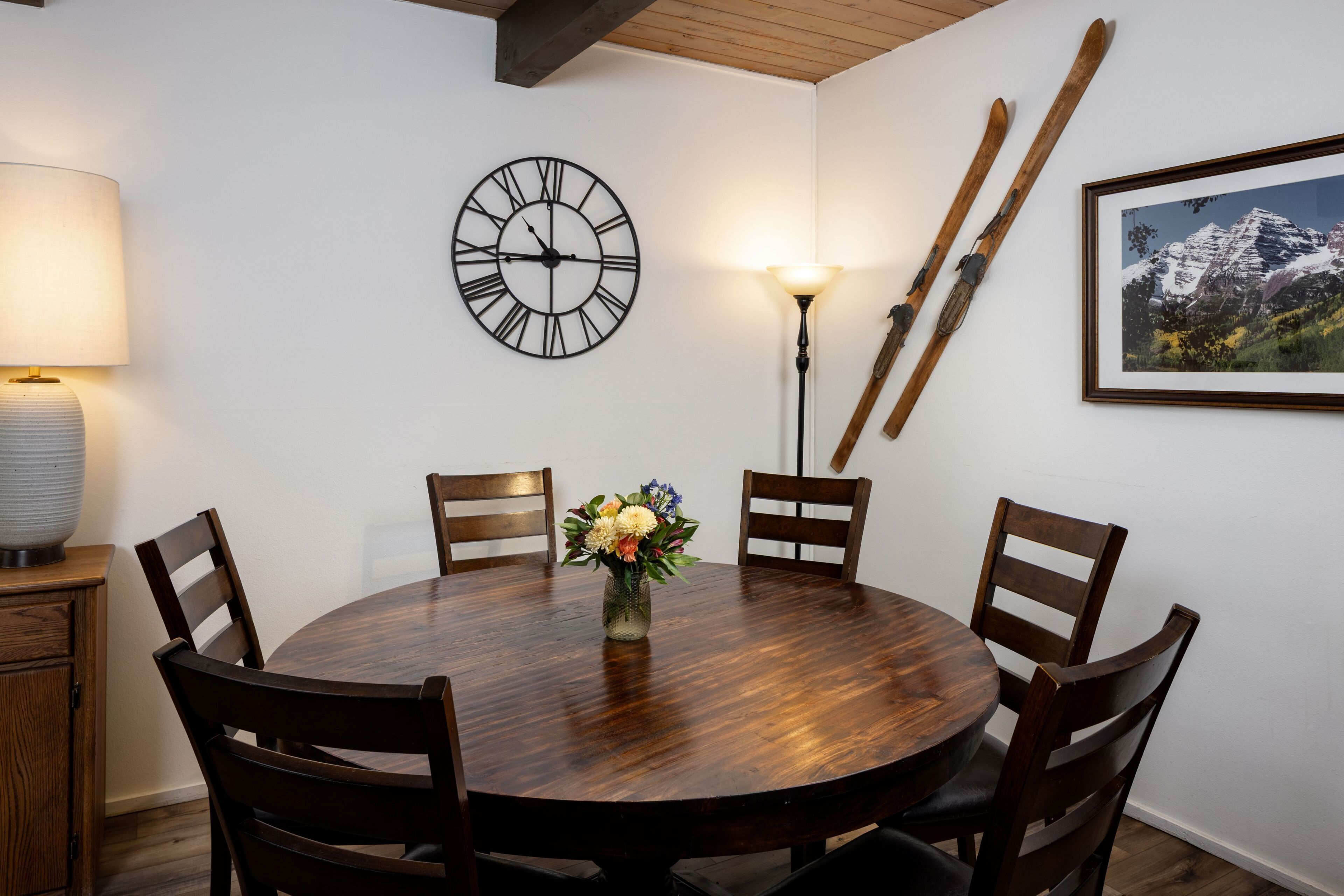 Uma mesa de jantar redonda de madeira com seis cadeiras, um relógio de parede, um abajur, um quadro emoldurado e esquis encostados na parede.