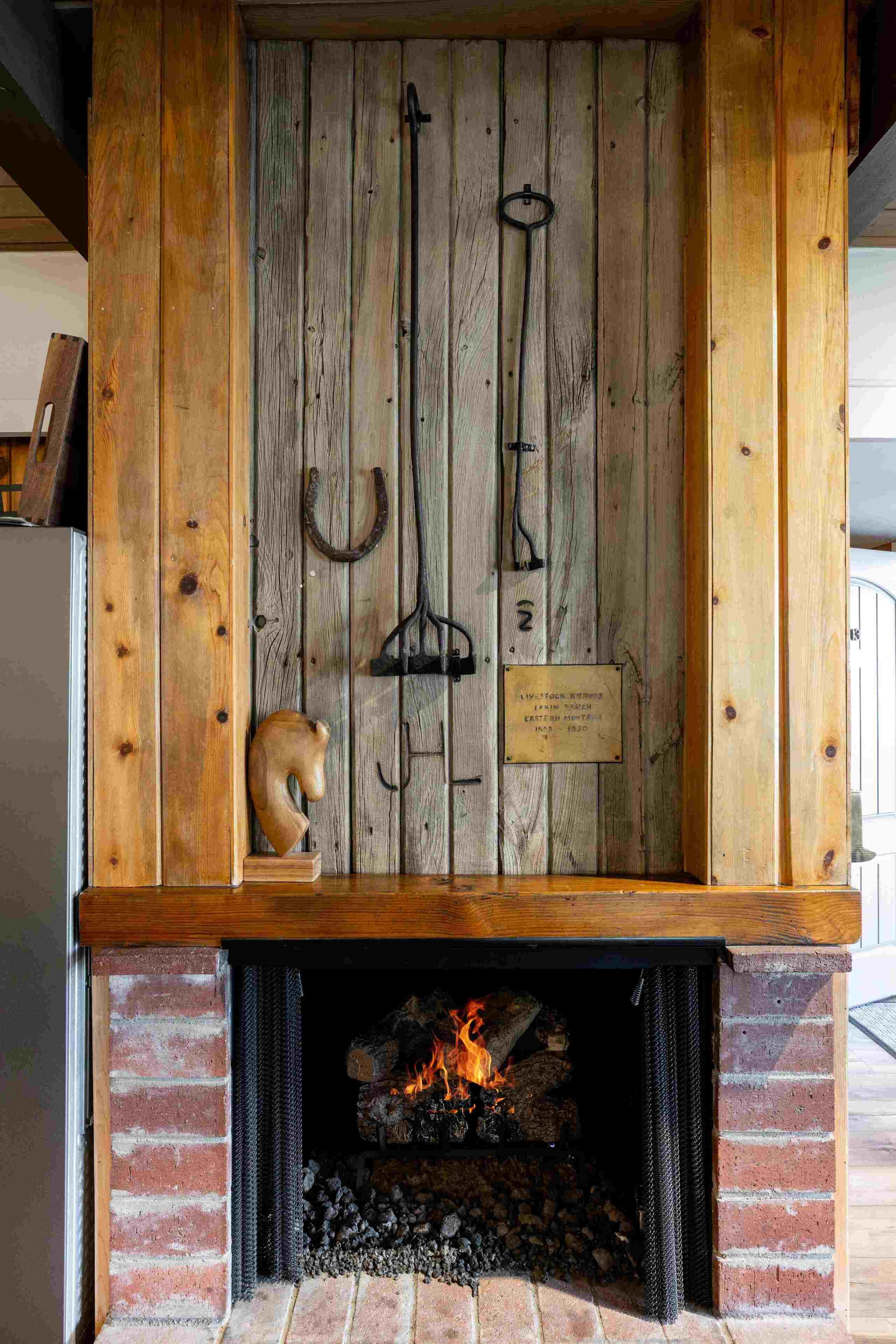 A fireplace with a wooden mantel and a plaque, a fire burning inside, and a rustic wall with hanging items.