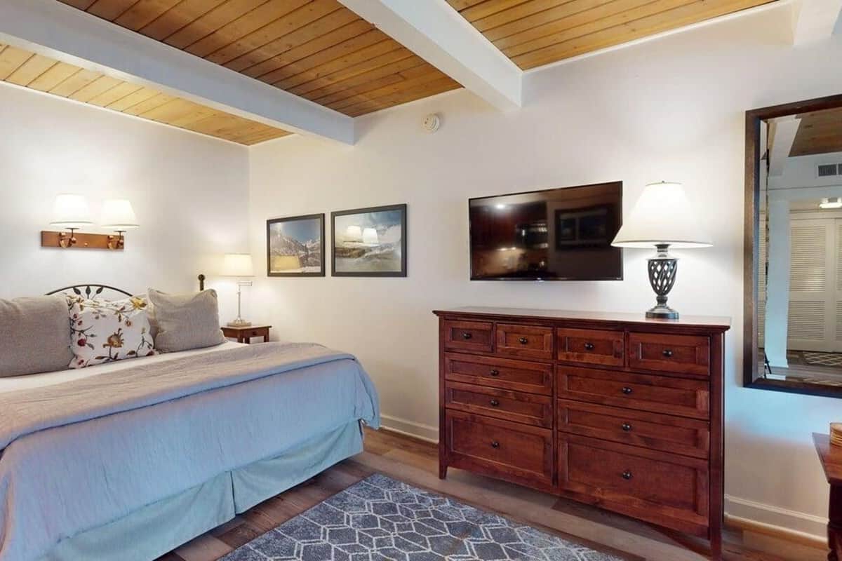 A bedroom with a bed, wooden dresser, wall-mounted TV, and a window with blinds.