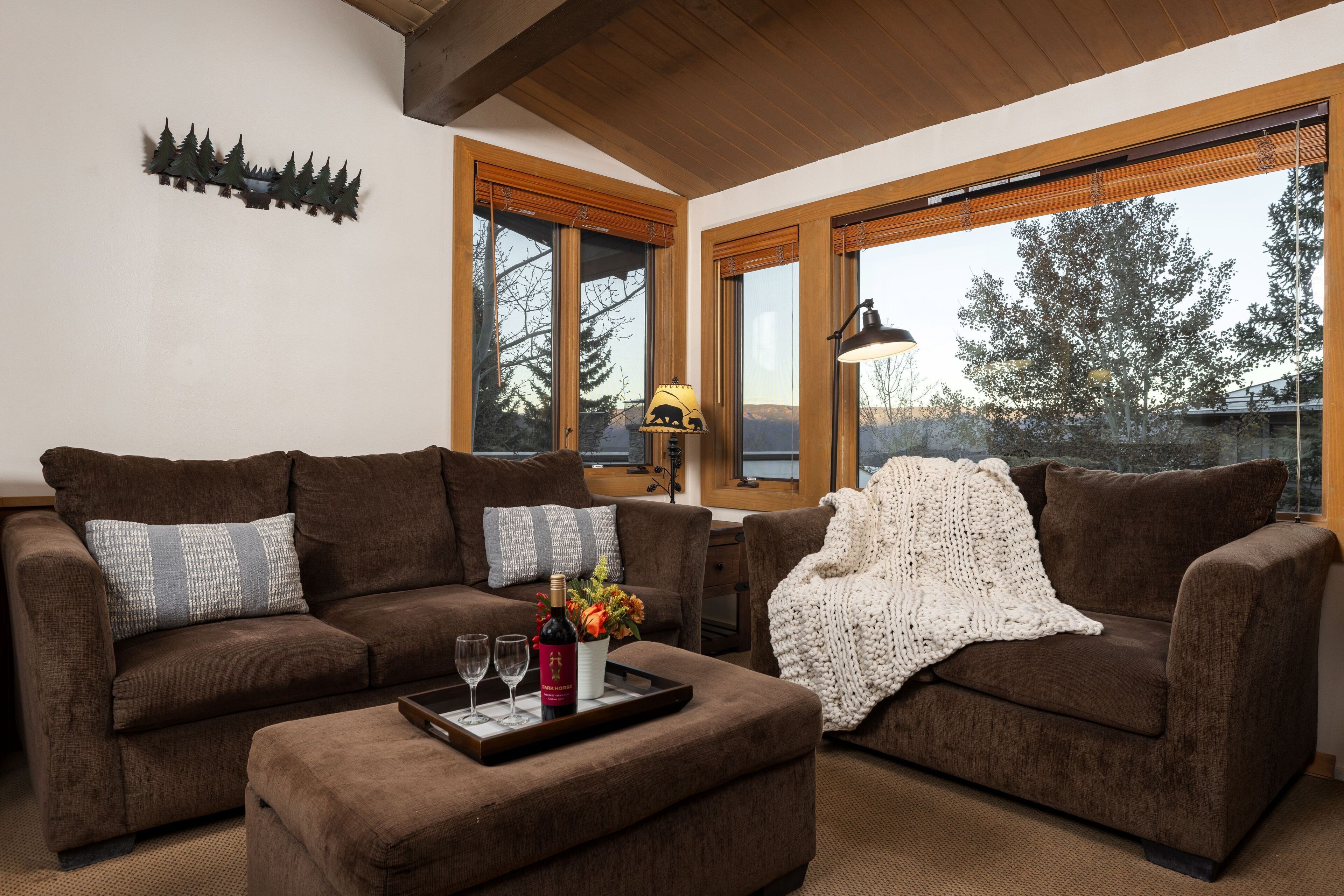 A cozy living room with two brown sofas, a coffee table with a bottle and glasses, and a large window with a view of trees.