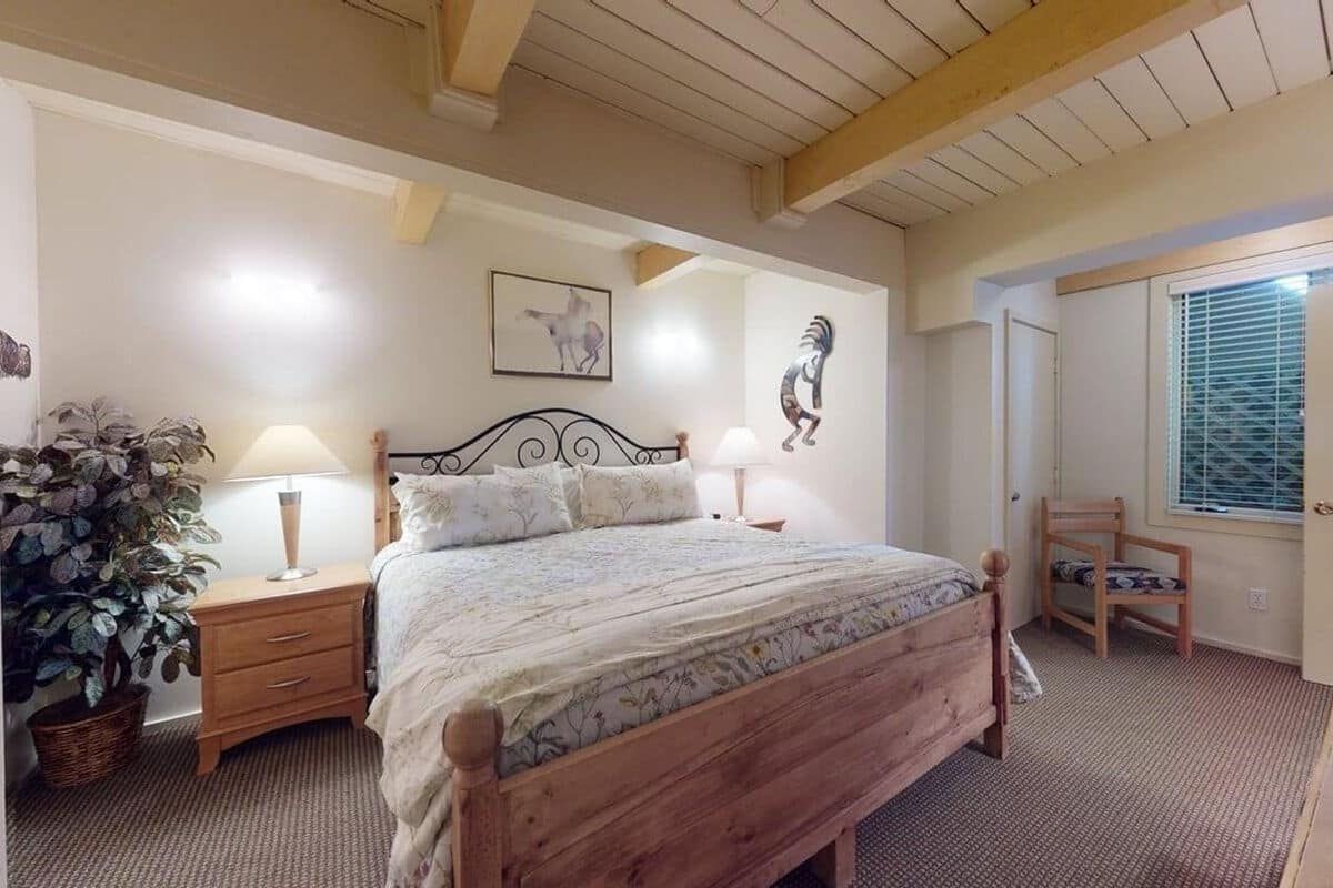 A bedroom with a wooden bed, two bedside tables with lamps, a wall-mounted artwork, a potted plant, and a window with blinds.