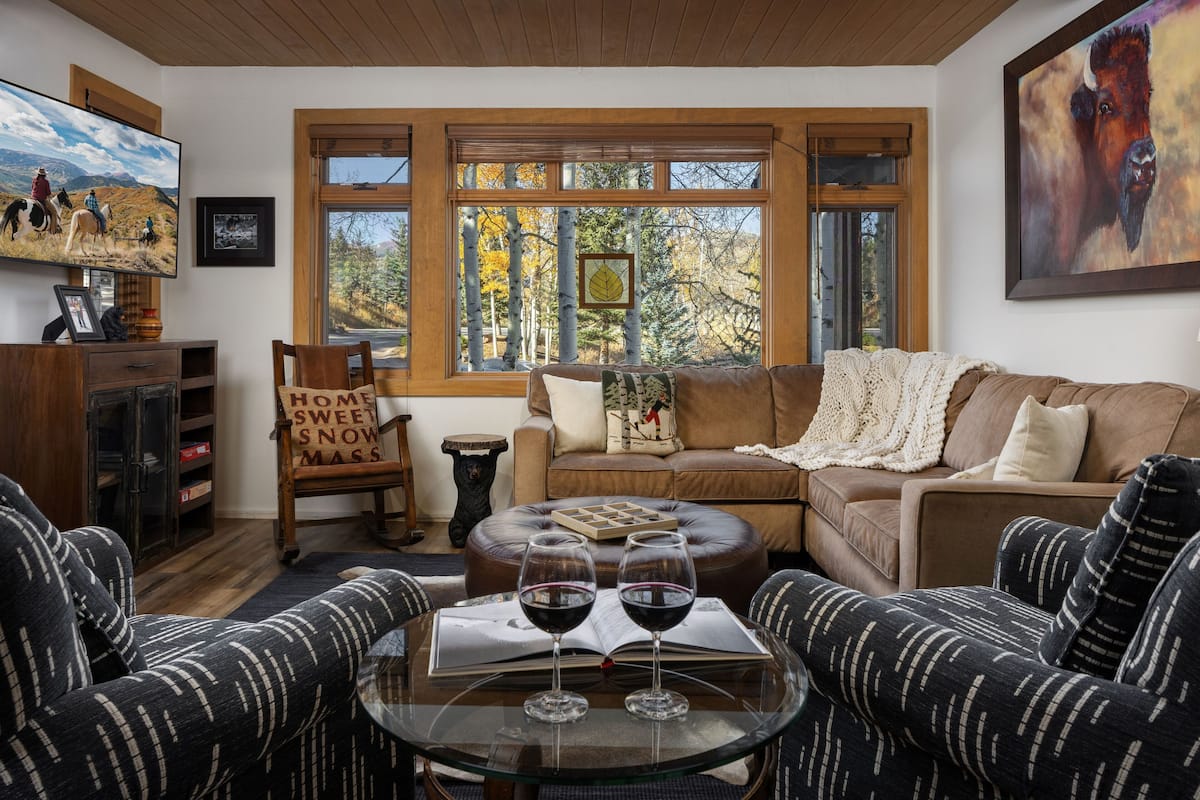 A cozy living room with a sectional sofa, a glass coffee table with two wine glasses, a television, and a large window with a view of trees.