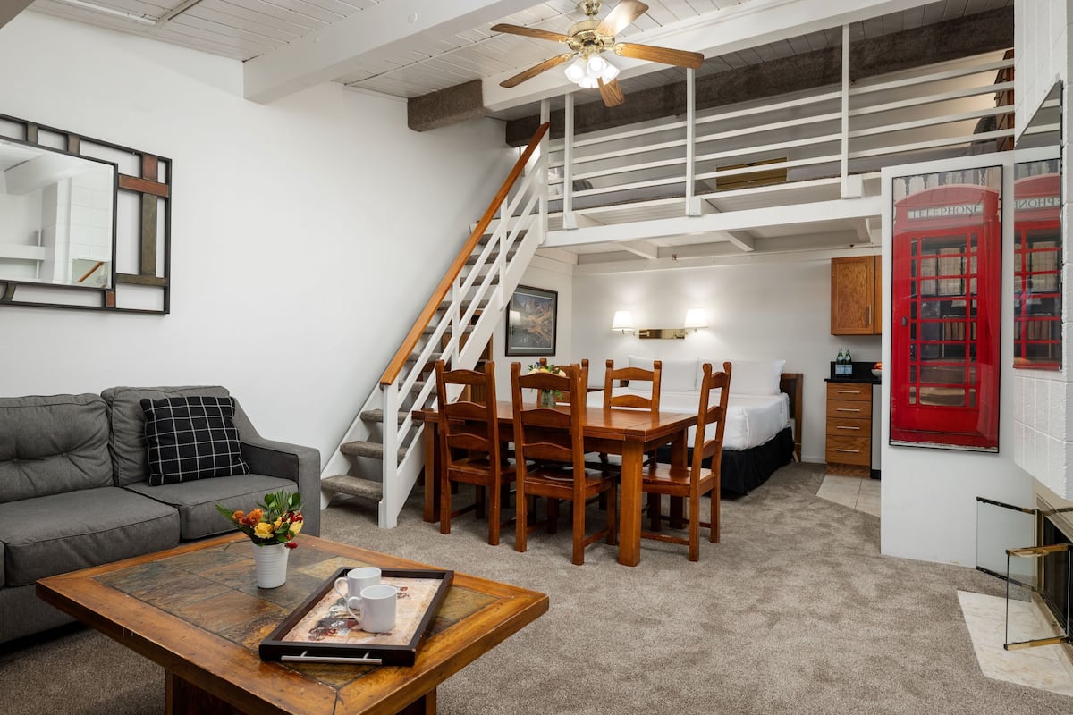 A living room with a sofa, coffee table, and a dining area with wooden chairs and a table. There is a staircase with a white railing leading to an upper level.