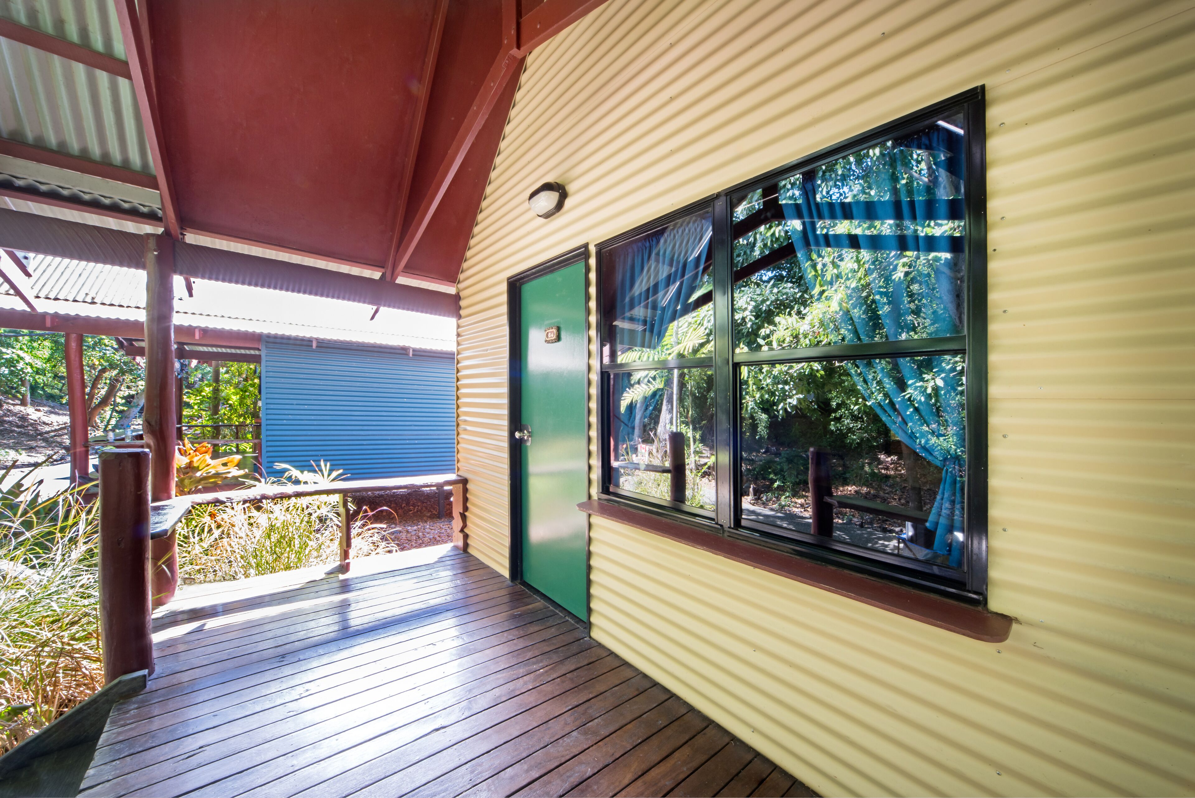 A covered wooden deck with a bench, a green door, and a window with blue curtains.