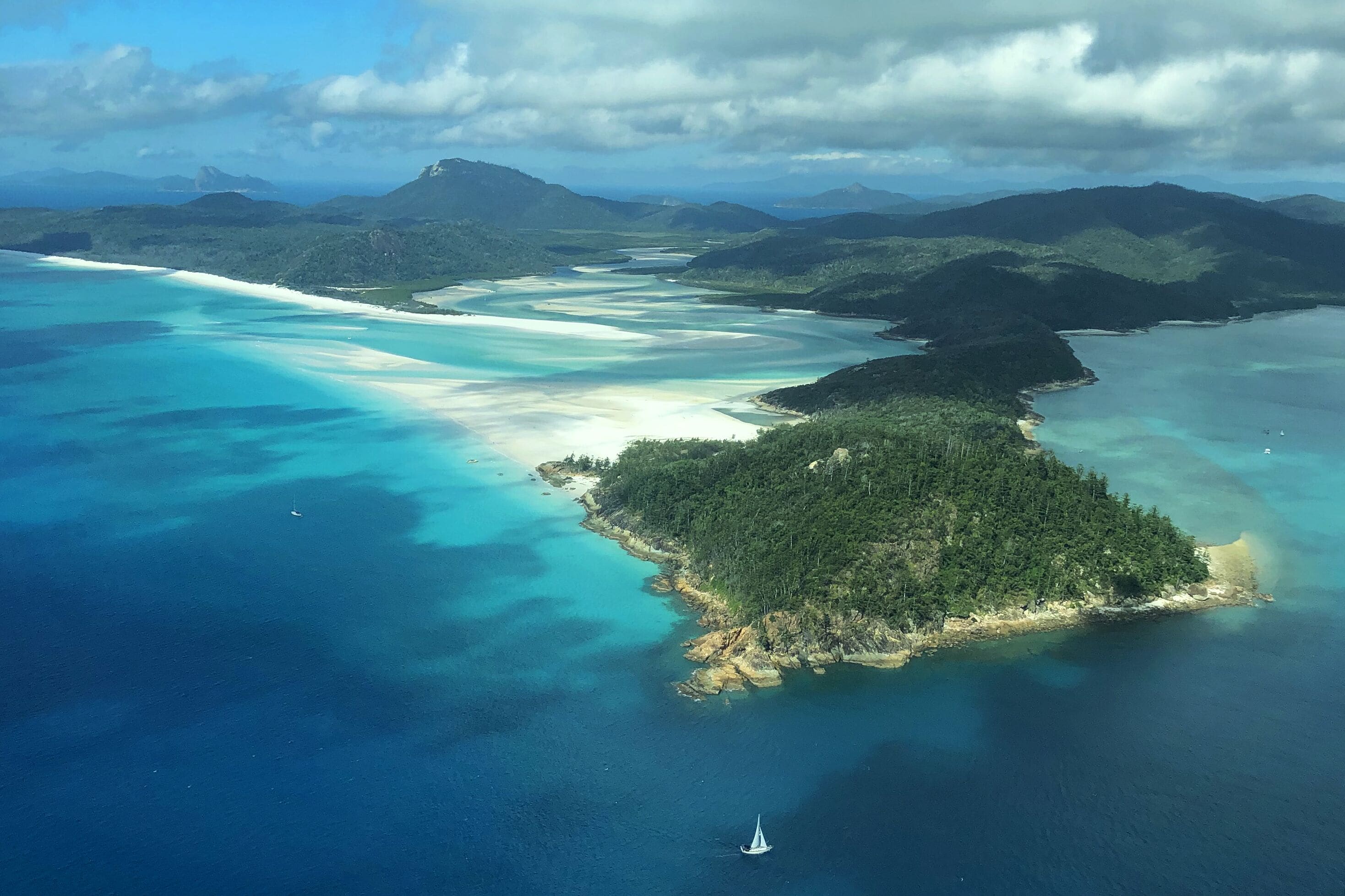 Une vue aérienne d’un paysage côtier, avec une petite île entourée d’eaux turquoise et une chaîne de montagnes lointaine.