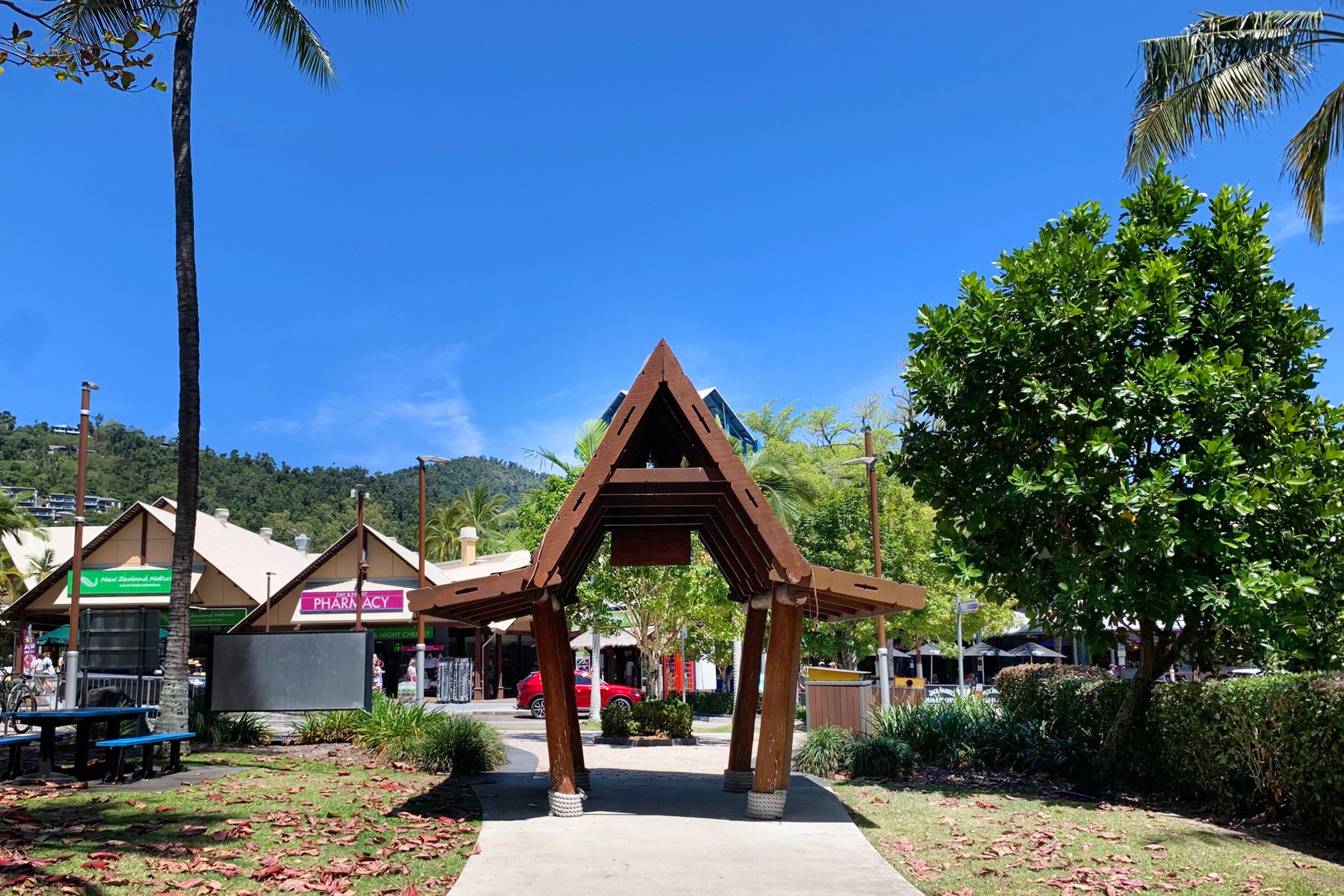 A wooden pavilion with a triangular roof structure, surrounded by greenery and a clear blue sky.
