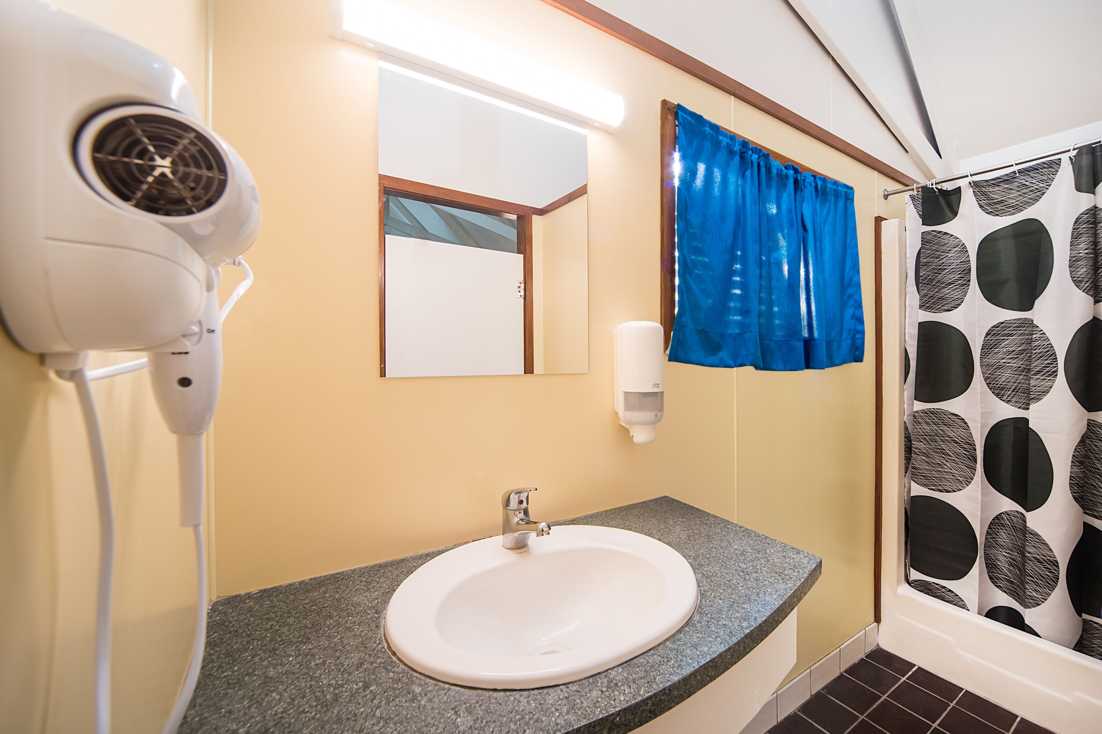 A bathroom with a white sink, a hairdryer, a mirror, a blue shower curtain, and a soap dispenser.