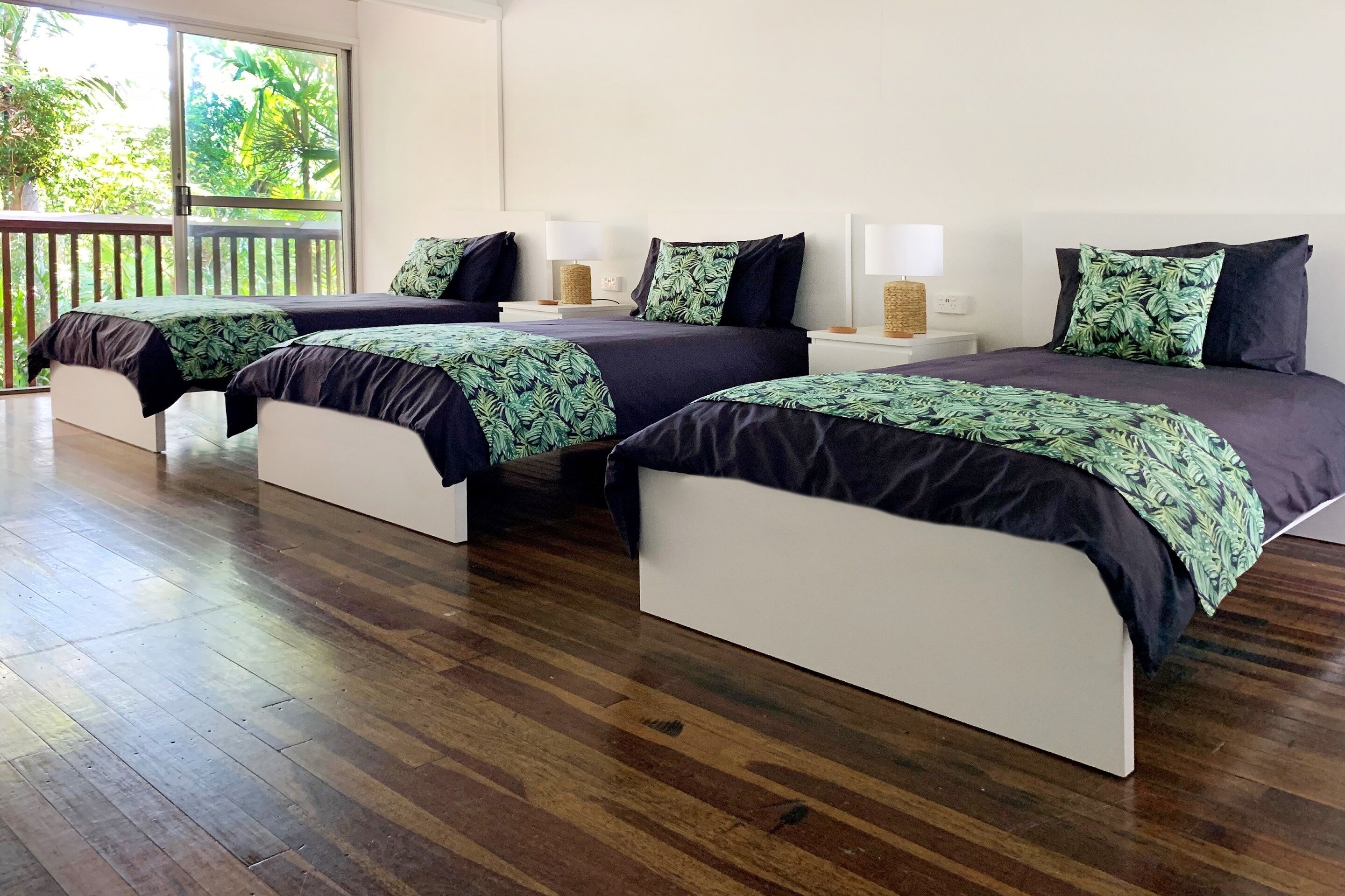 A hotel room with three beds, each with a dark-colored headboard and a green leaf-patterned bedspread. The room has a wooden floor and a large window providing a view of greenery outside.