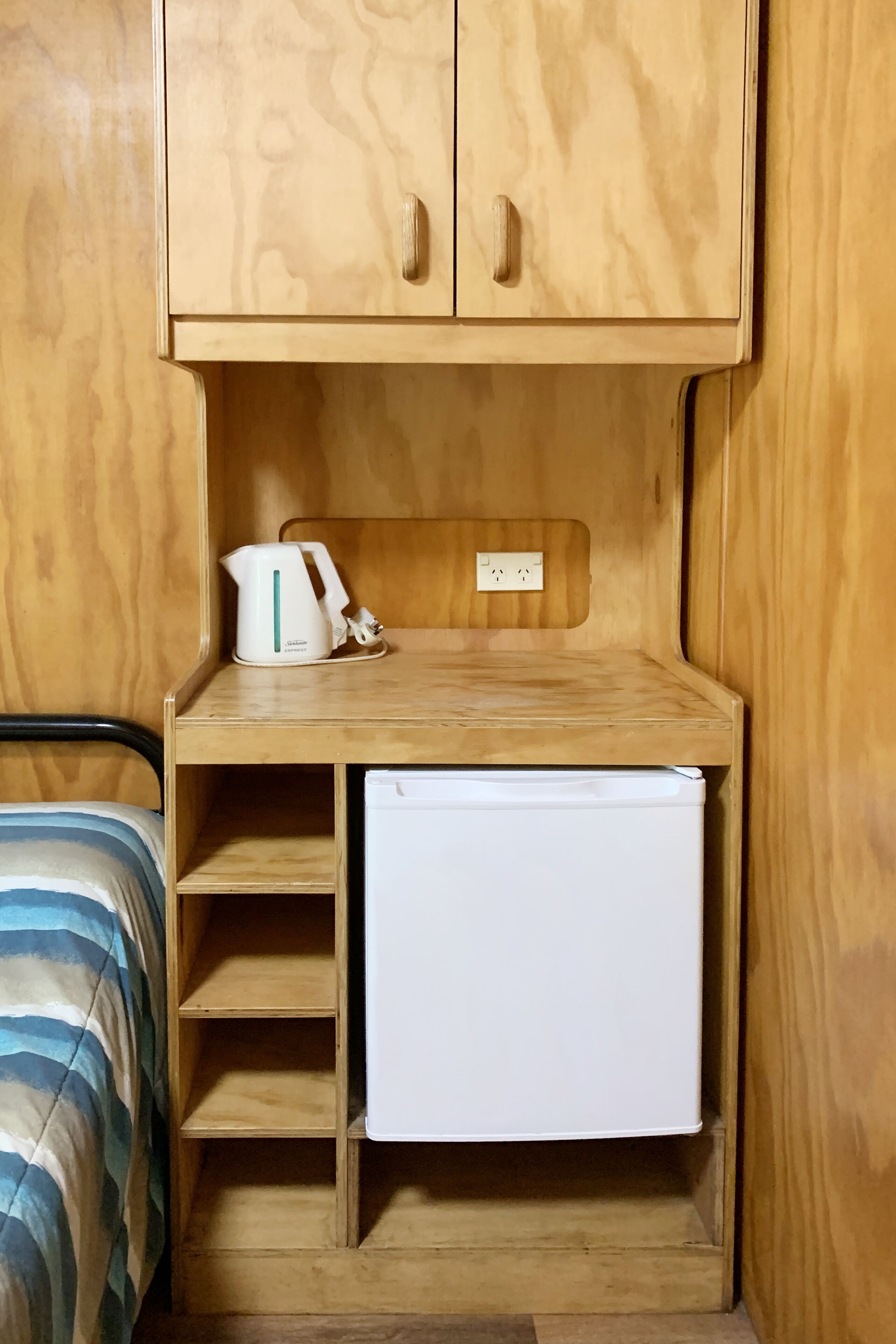 Una habitación compacta con un armario de madera, un refrigerador blanco pequeño y una cama con ropa de cama a rayas.