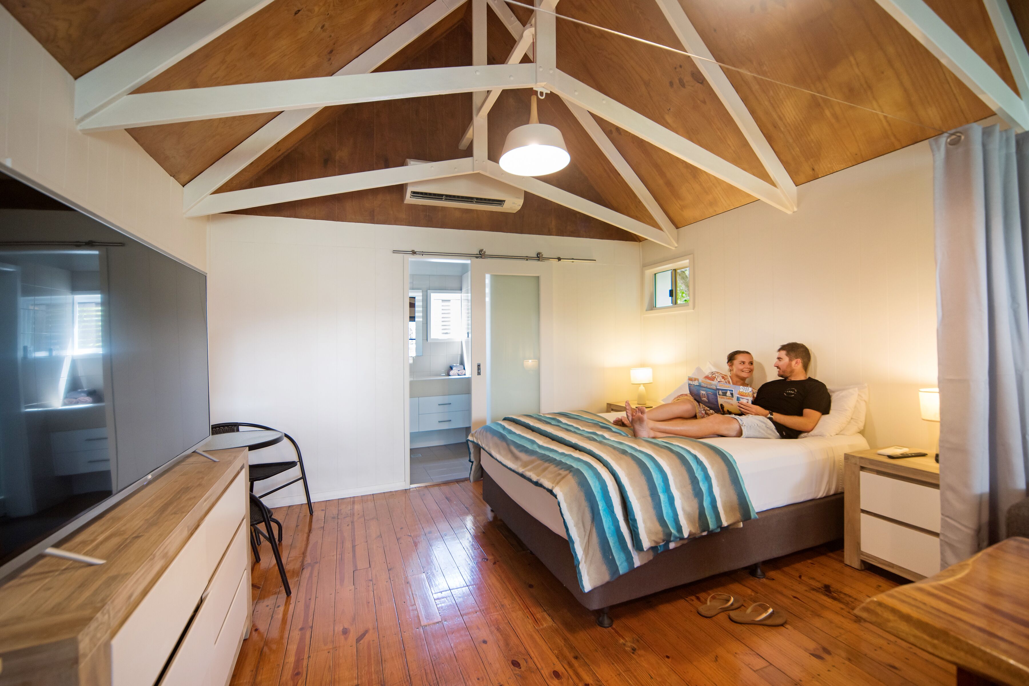 A bedroom with a wooden ceiling, a bed with striped bedding, a bedside table, and a nightstand.