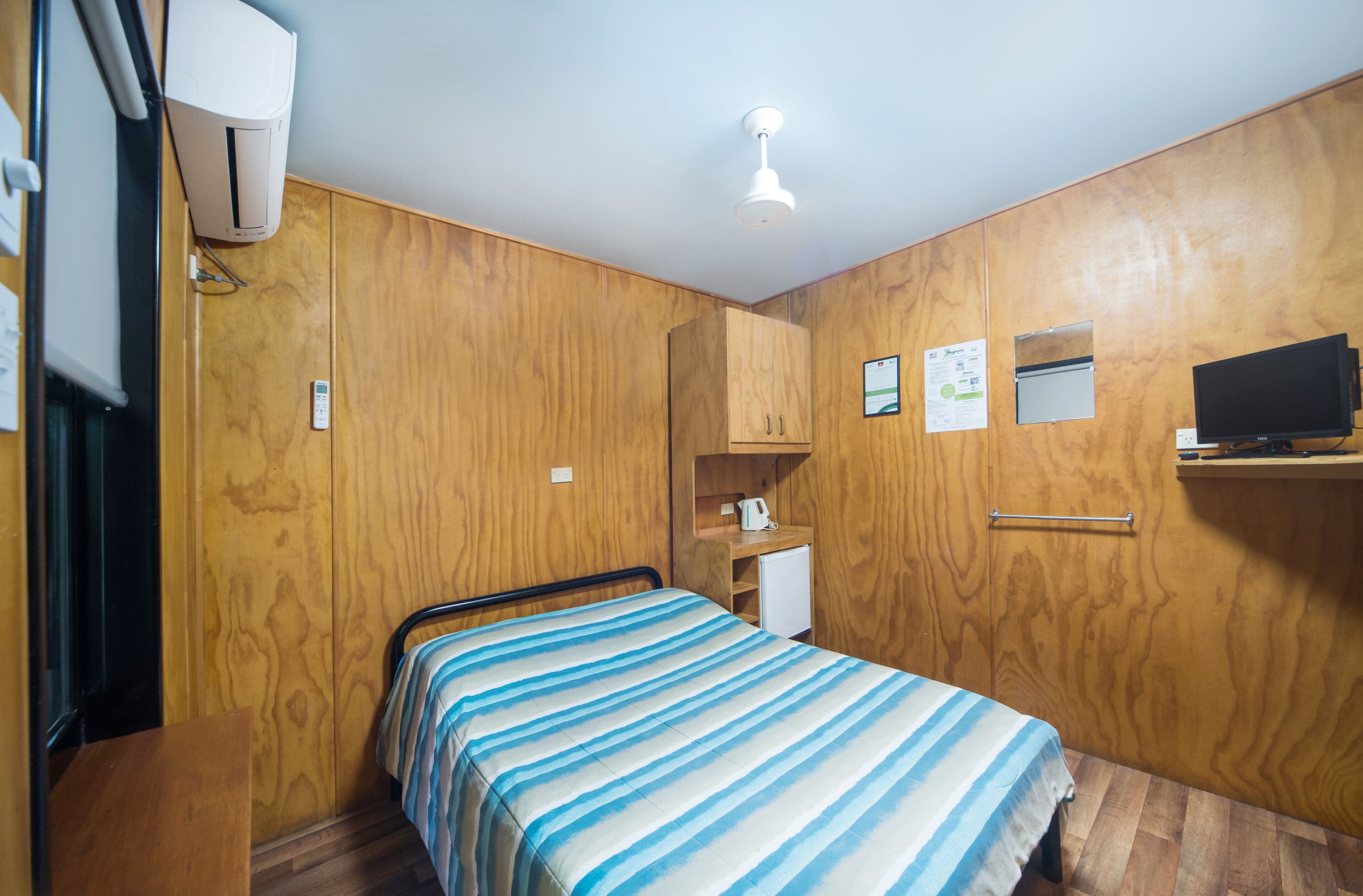 Habitació compacta amb llit, parets de fusta, un escriptori petit, televisió i ventilador de sostre.