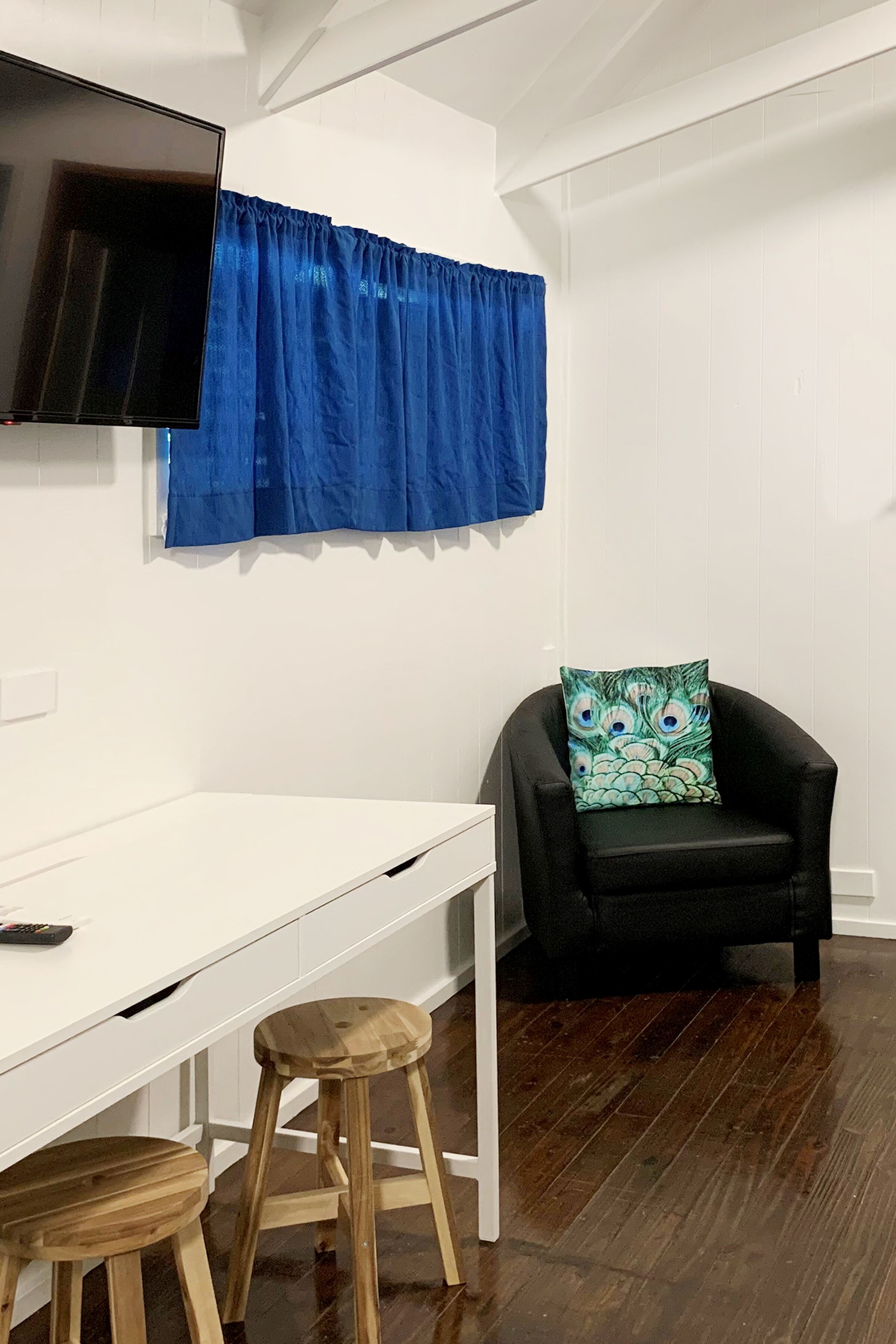Une pièce avec un bureau blanc, deux tabourets en bois, une chaise noire avec un coussin à motifs, un rideau bleu et un téléviseur à écran plat fixé au mur.