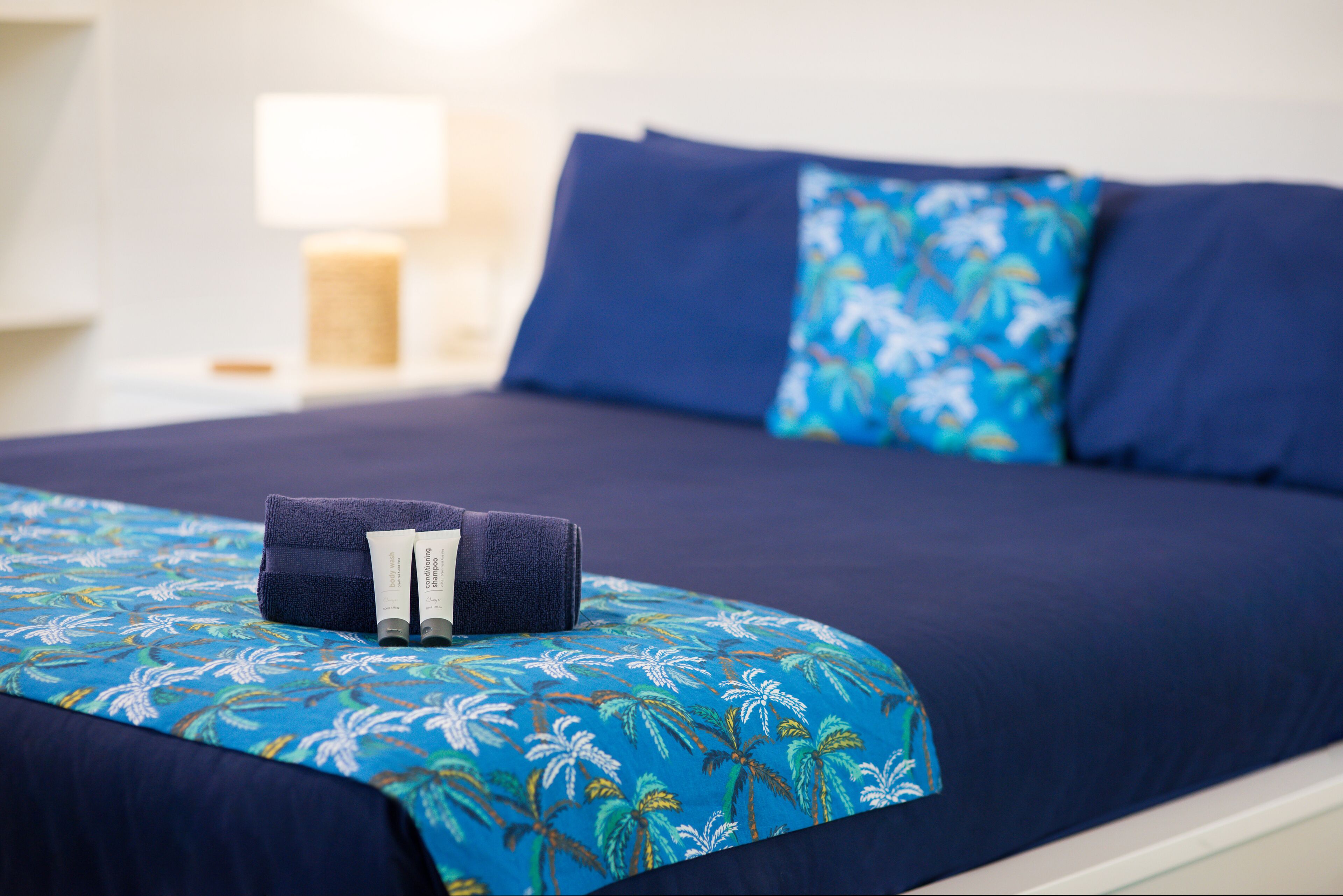 A neatly made bed with a blue floral bedspread, a dark blue pillow, and a matching pillow on a nightstand with a lamp.