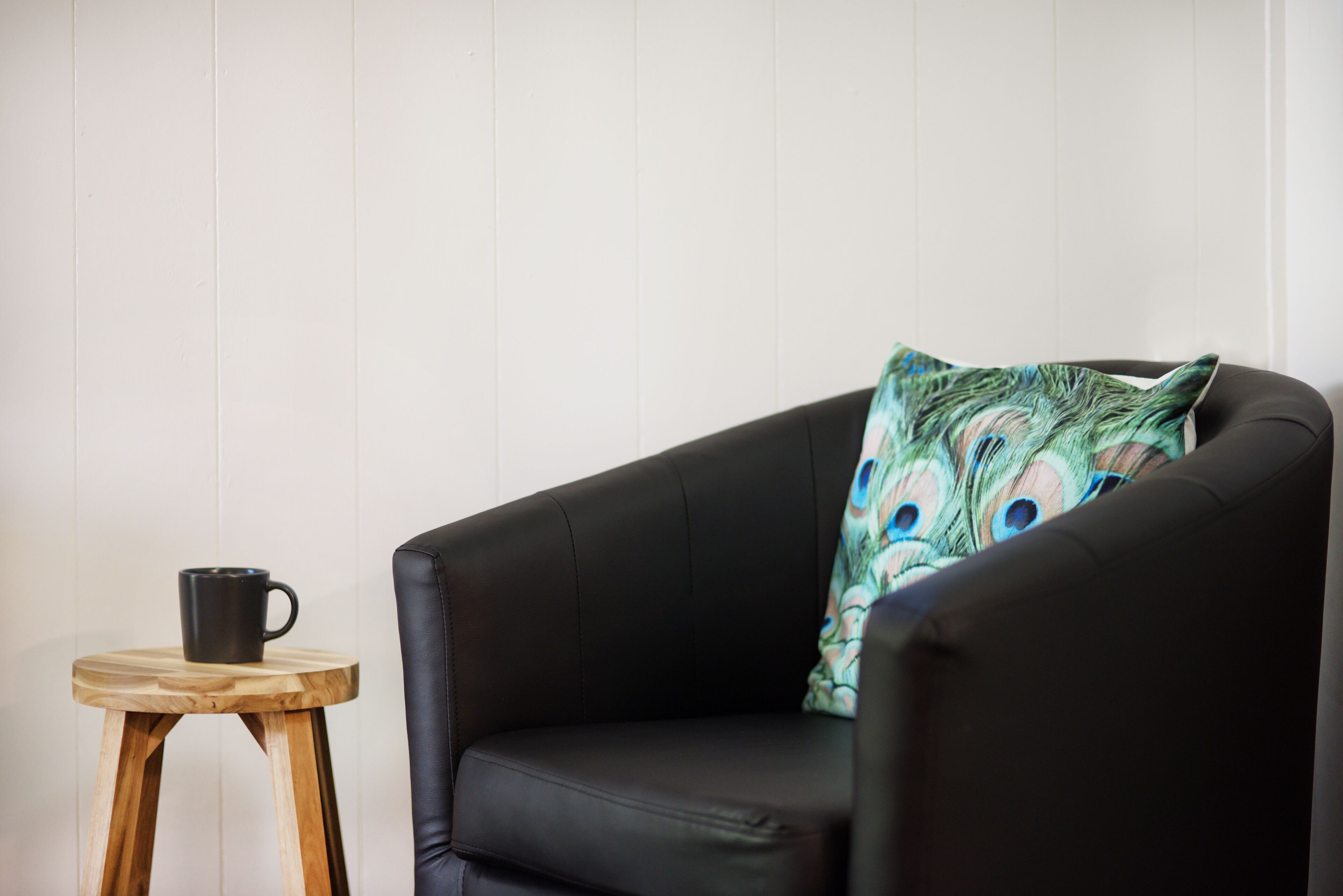 Un fauteuil en cuir noir agrémenté d’un coussin orné d’une plume de paon, un tabouret en bois avec une tasse noire, et des murs blancs à panneaux.