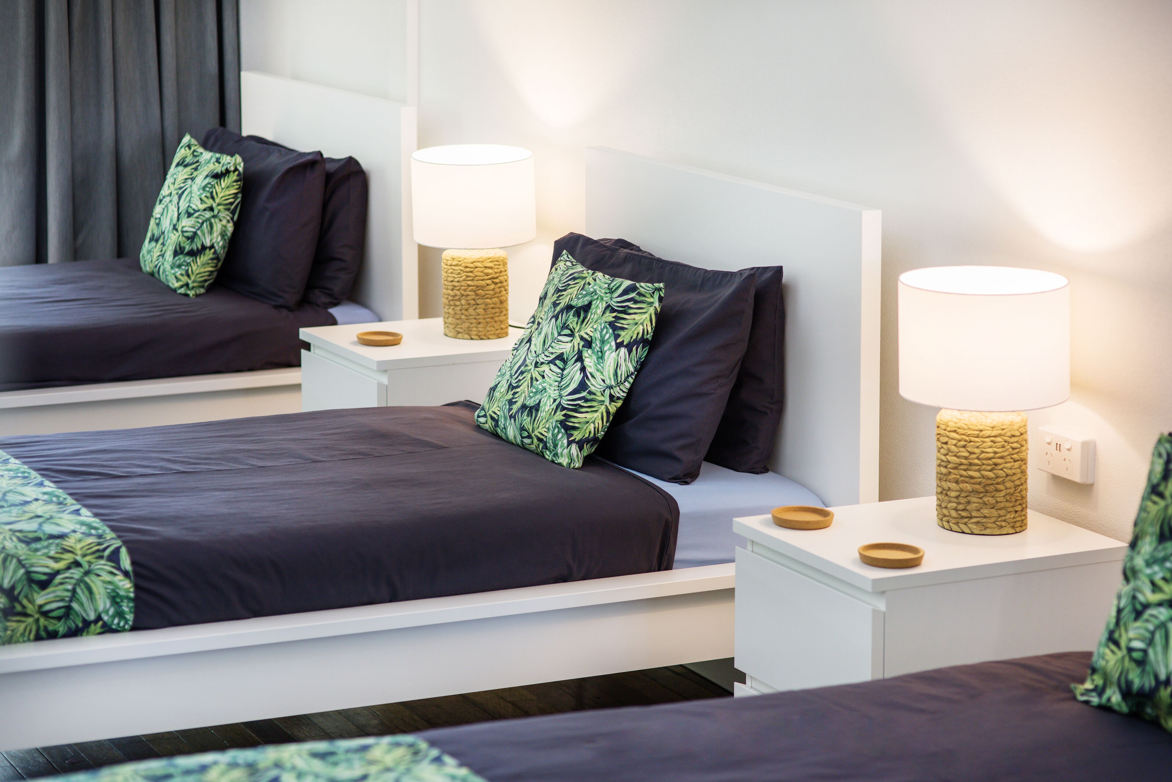 A hotel room with two beds, each with a dark-colored bedspread and green leaf-patterned pillows. There are bedside tables with lamps and a white headboard.