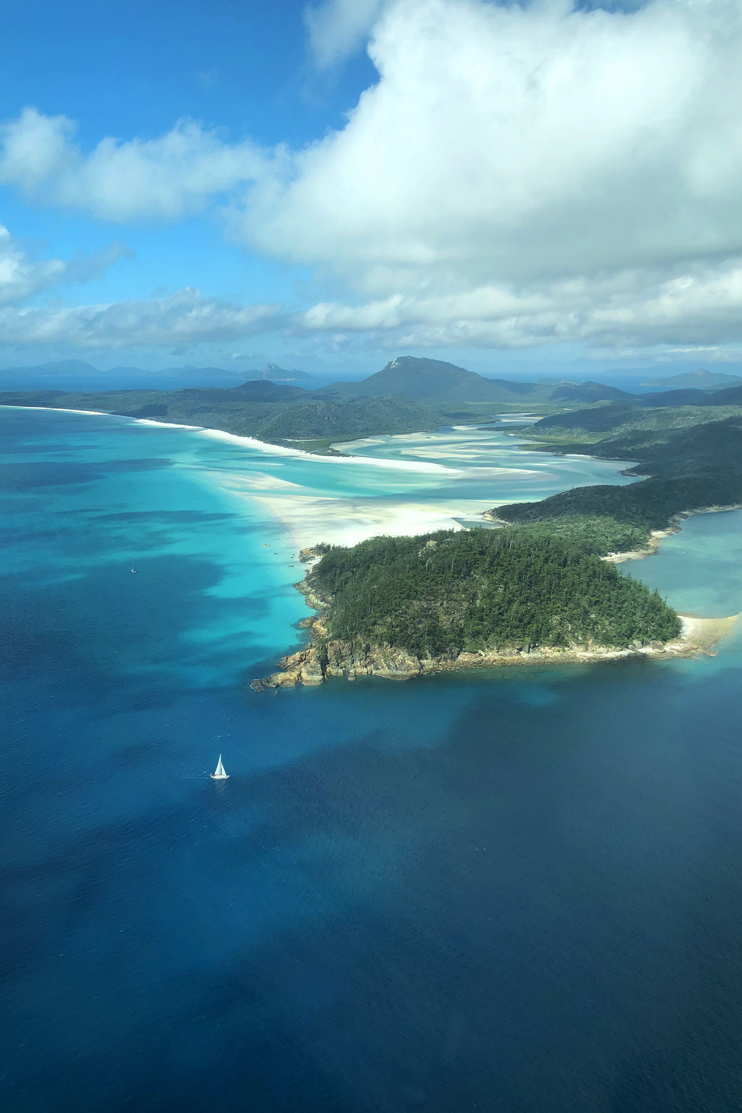 An aerial view of a coastal area with clear blue waters, a small island with dense greenery, and distant mountains under a partly cloudy sky.
