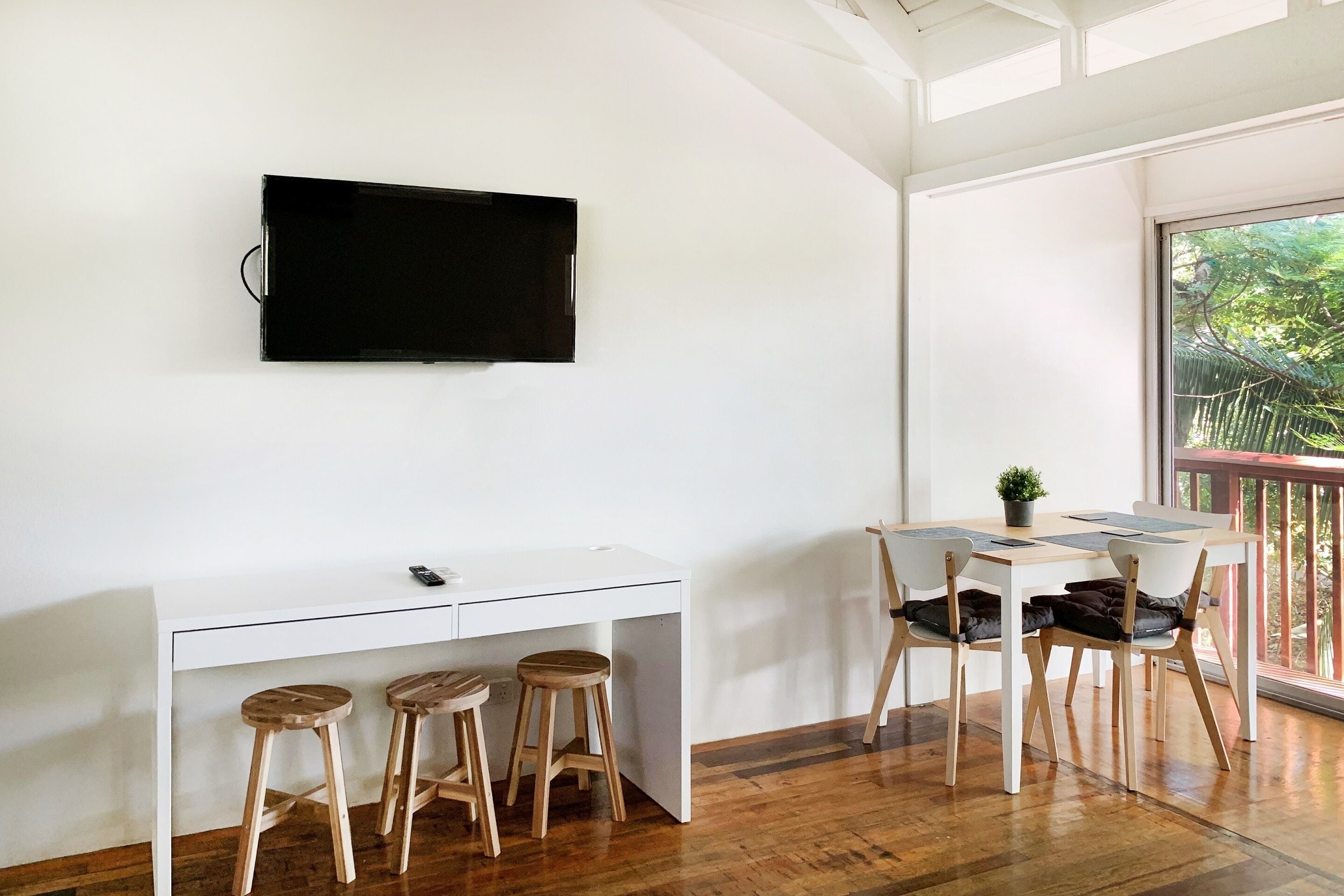 Una habitación moderna con un escritorio blanco, dos taburetes de madera, un televisor de pantalla plana en la pared y un balcón con vistas a zonas verdes.