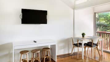Una habitación moderna con un escritorio blanco, dos taburetes de madera, un televisor de pantalla plana en la pared y un balcón con vistas a zonas verdes.