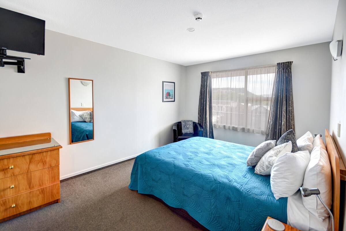 A bedroom with a bed, a dresser, a chair, a TV mounted on the wall, and a window with curtains.