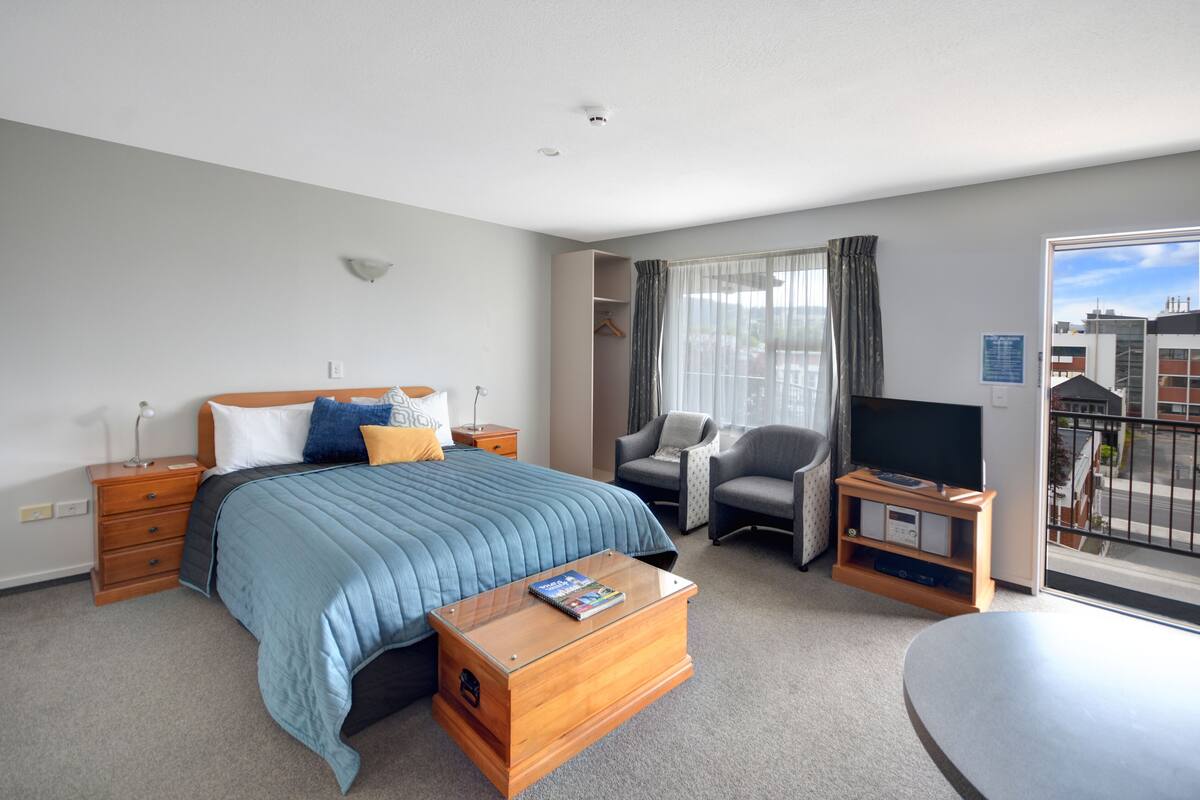 A modern hotel room with a large bed, a desk, a chair, a TV, and a balcony with a city view.