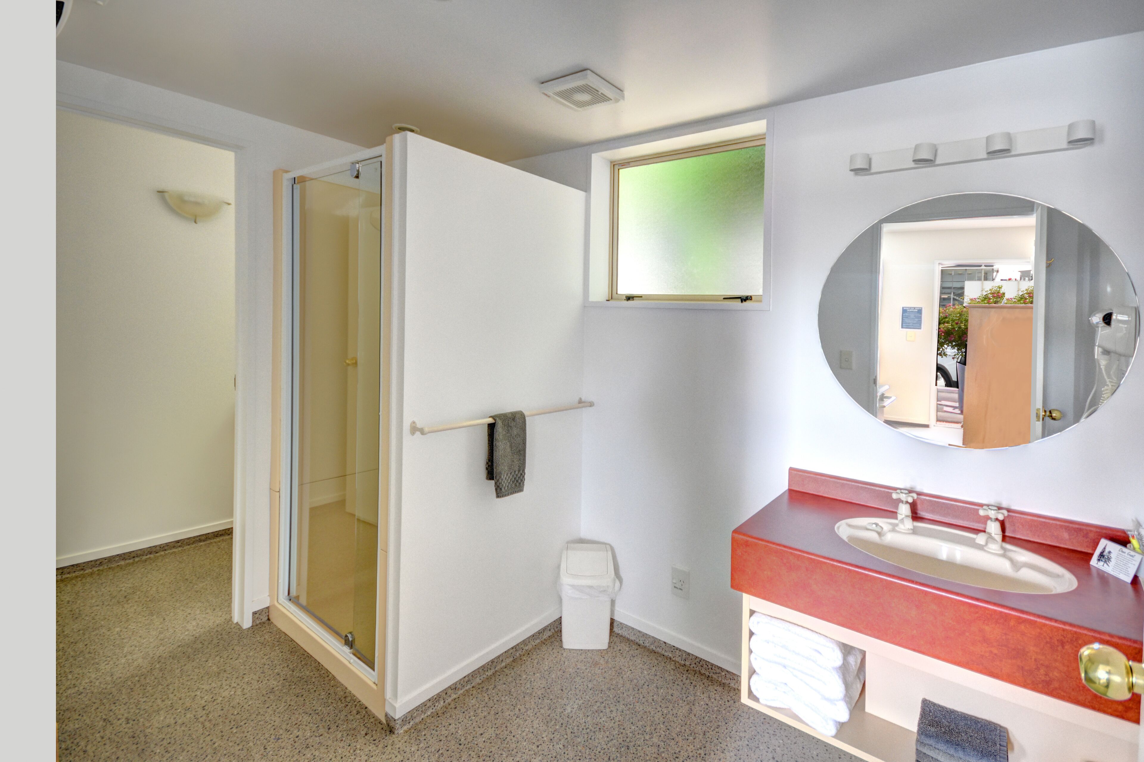 Une salle de bain avec une porte de douche en verre, un lavabo avec un plan de toilette rouge, un miroir et un porte-serviettes.