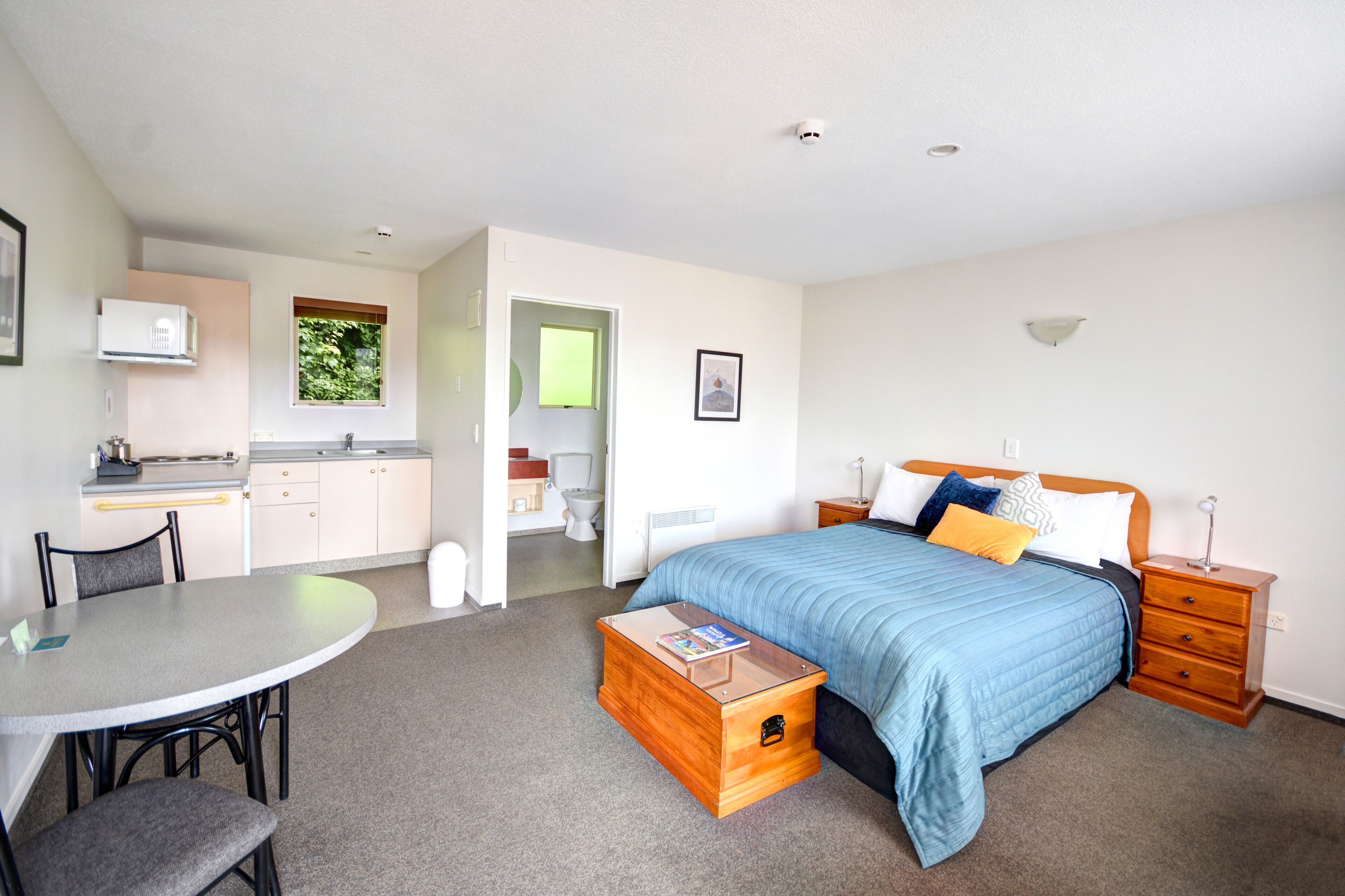 A compact hotel room with a bed, bedside table, and a small wooden chest of drawers. A kitchenette with a microwave and a window is visible in the background.