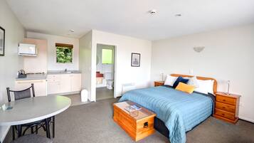 A compact hotel room with a bed, bedside table, and a small wooden chest of drawers. A kitchenette with a microwave and a window is visible in the background.