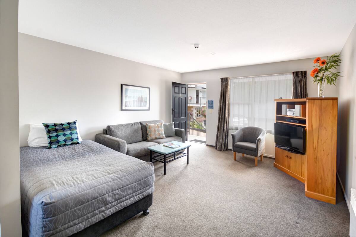 A modern hotel room with a large bed, a sofa, a coffee table, a TV stand, and a balcony with curtains.